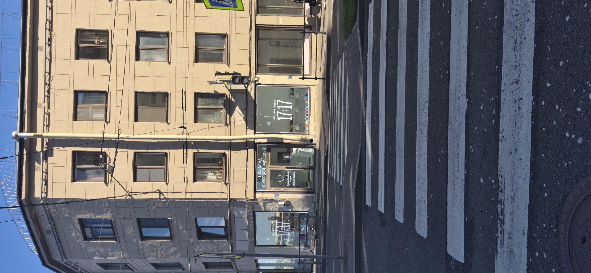 The image shows a street scene in Saint Petersburg, Russia, with a modern building facade and a crosswalk in the foreground. The building is a light beige color with multiple windows. On the ground floor, there are storefronts. One storefront has the Cyrillic text "ШОКОЛАД С ОСОБОЙ МИССИЕЙ" (Chocolate with a Special Mission) above its entrance, along with a heart symbol and smaller text that appears to be "COFFEE, HOT CHOCOLATE". Another storefront to the right has large numbers "17:17" displayed prominently on its window, with the Cyrillic text "ВЕРЬ, МЕЧТА СБЫВАЕТСЯ" (Believe, a dream comes true) above it. A traffic light is visible on a pole attached to the building, and a pedestrian crossing (crosswalk) is painted on the asphalt street in the foreground. The sky is clear and blue, indicating daytime. Electrical wires are visible strung across the facade of the building.