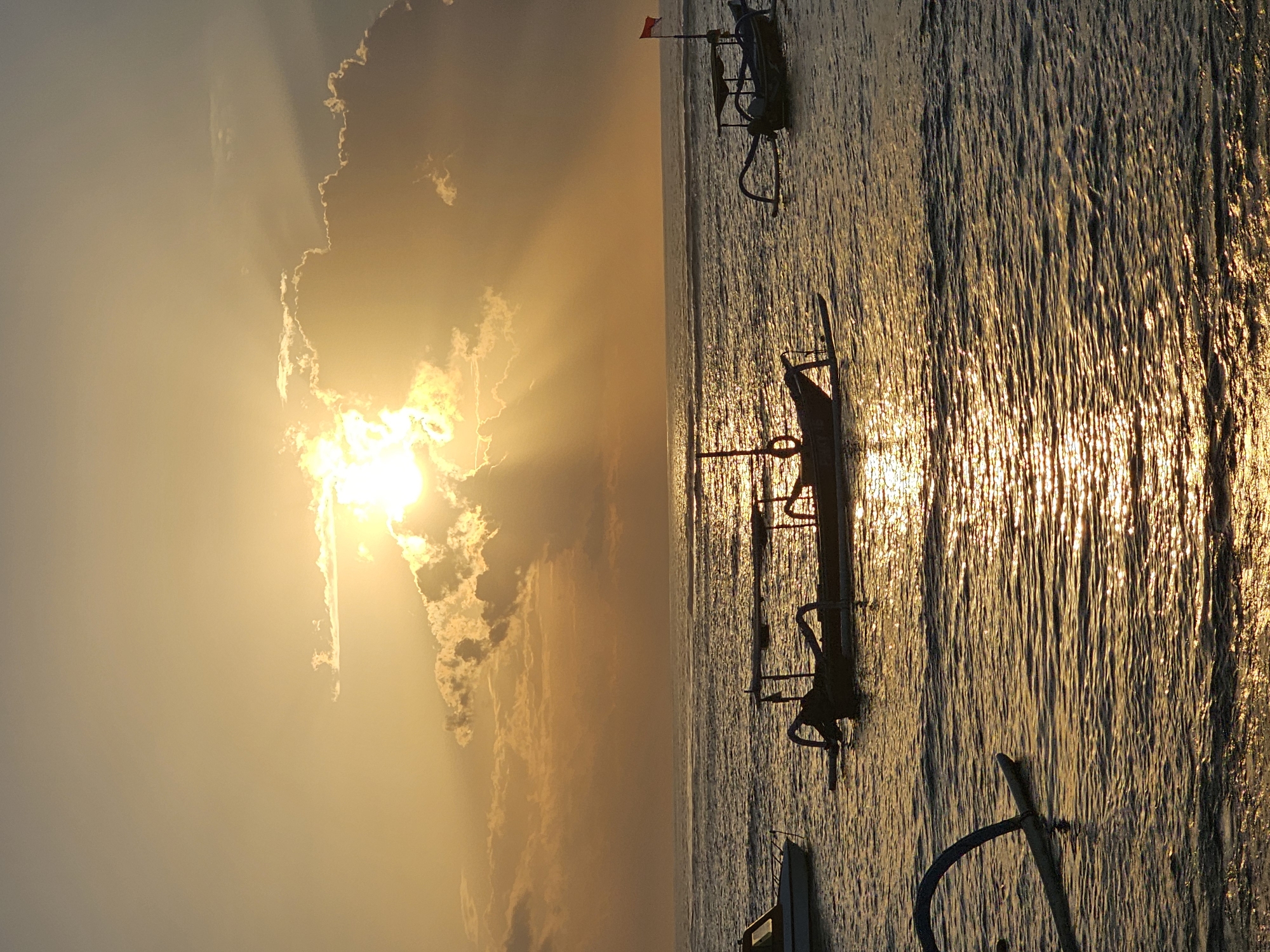 This is a vertical, medium shot of a sunset over the ocean in Denpasar, Indonesia. The sun is partially obscured by clouds, casting golden rays across the water. Three boats are visible on the water: a white boat on the left, a traditional outrigger canoe in the center, and another outrigger canoe with a red flag on the right. The water is calm with gentle ripples, reflecting the warm hues of the sunset. The sky is a gradient of yellow and orange, transitioning to a darker tone near the top of the frame. The image captures a serene and peaceful moment at the end of the day.