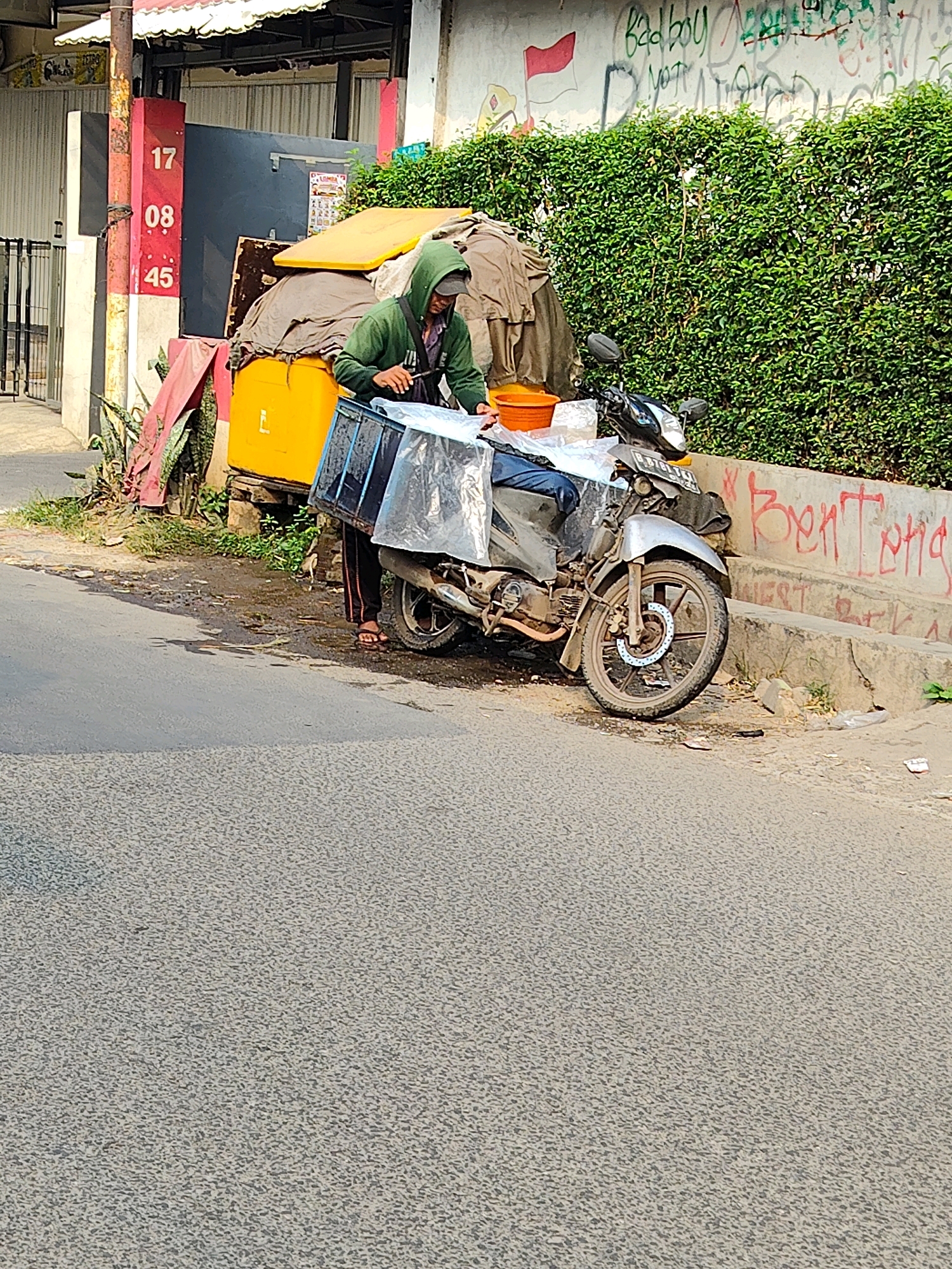 A person wearing a green hooded sweatshirt is standing next to a motorcycle on the side of a street in Bekasi, Indonesia. The motorcycle has a clear plastic cover over a cargo box attached to the rear. The person appears to be interacting with the cargo box. Behind the motorcycle, there is a bright yellow container and a wall with red graffiti that reads "Benteng". A dense green hedge runs along the top of the wall. In the foreground, the street is paved with asphalt, and there are some puddles and debris on the ground. To the left, a red and yellow striped post with numbers "17", "08", and "45" is visible, along with a metal fence. The overall scene suggests an urban environment with some signs of commercial activity or daily life.