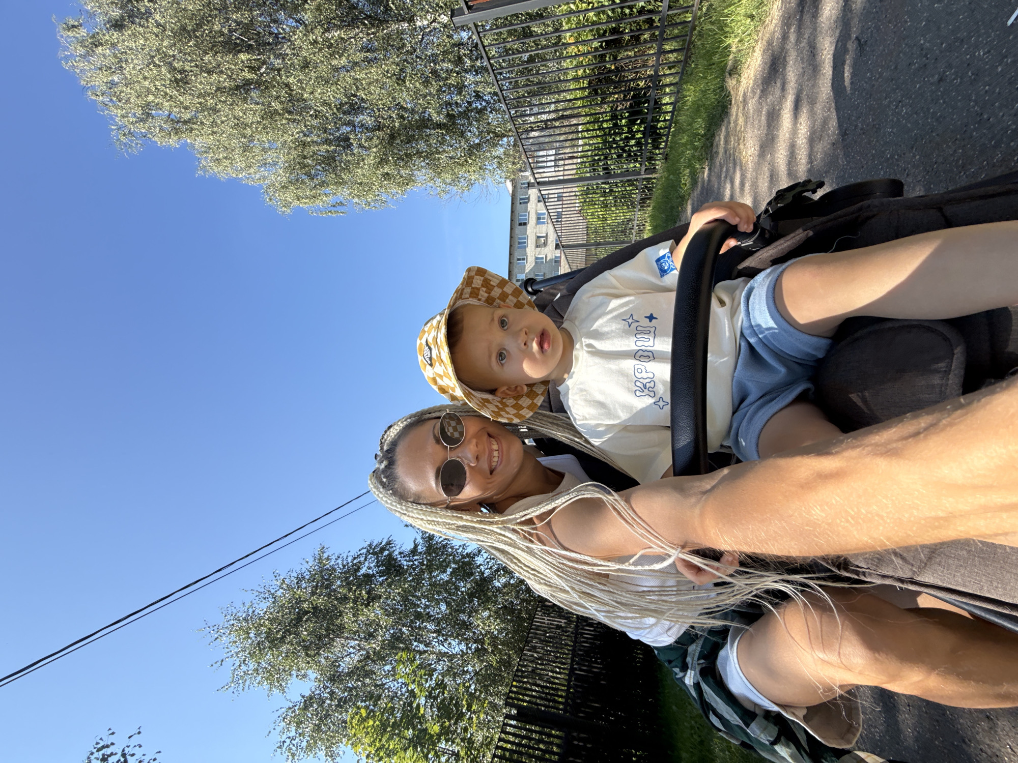 In Rusanovo, Russia, on a sunny day, a woman with blonde braided hair and sunglasses is taking a selfie in a stroller with a young child. The woman is wearing a white tank top and a green and white plaid shirt. The child is wearing a yellow checkered bucket hat and a white t-shirt with a blue graphic. They are on a paved path next to a black metal fence and trees. In the background, there is a residential building. The scene appears to be a park or a residential area.