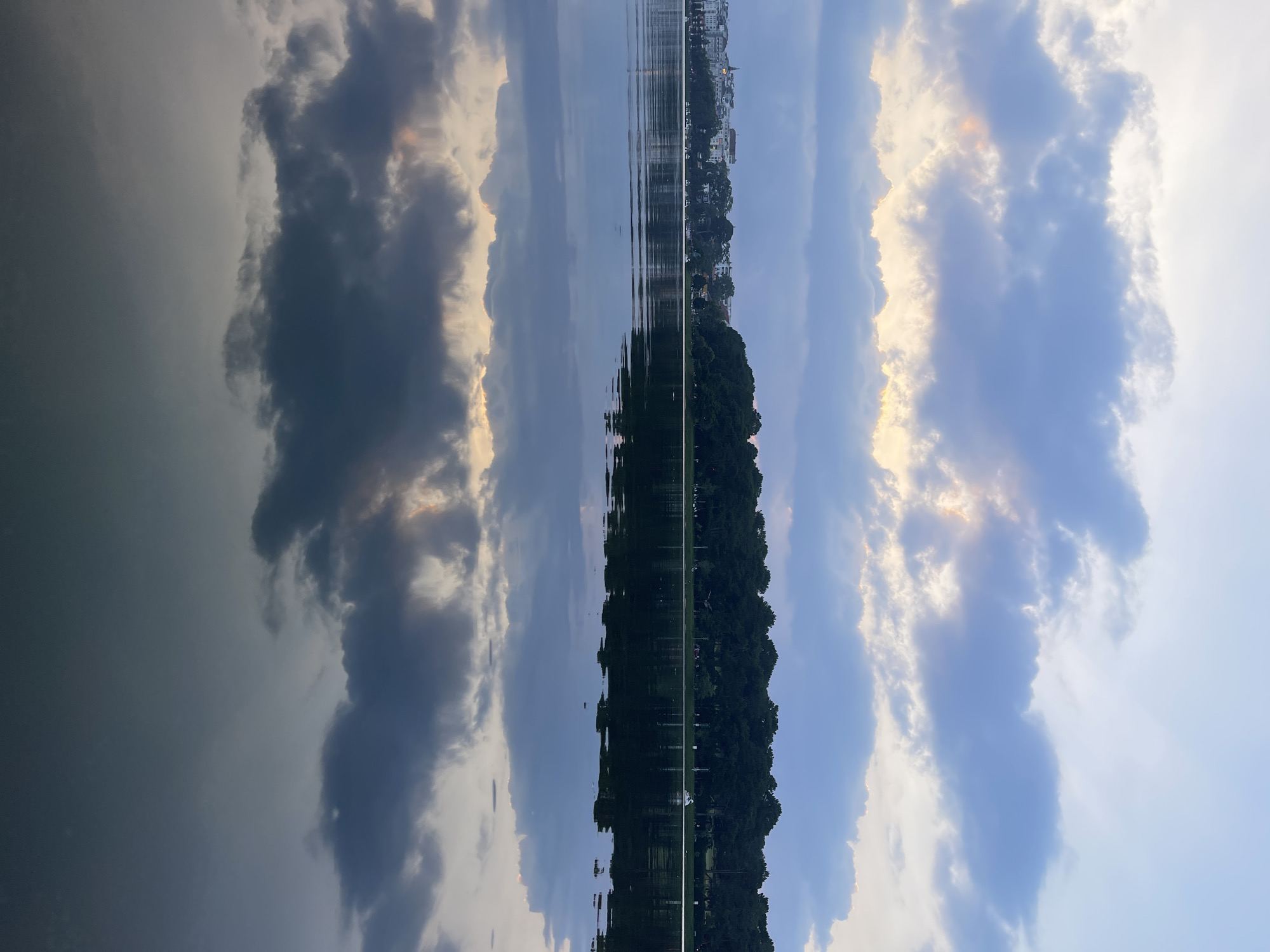 The image shows a symmetrical reflection in water, with an island of lush green trees in the center. On the upper portion of the image, building silhouettes are visible. The sky is filled with white and blue clouds with golden edges, mirrored on the other side of the trees, as well as reflected in the water. This location is likely in Da Lat, Vietnam, given the prevalent pine forests of that area and the urban setting.