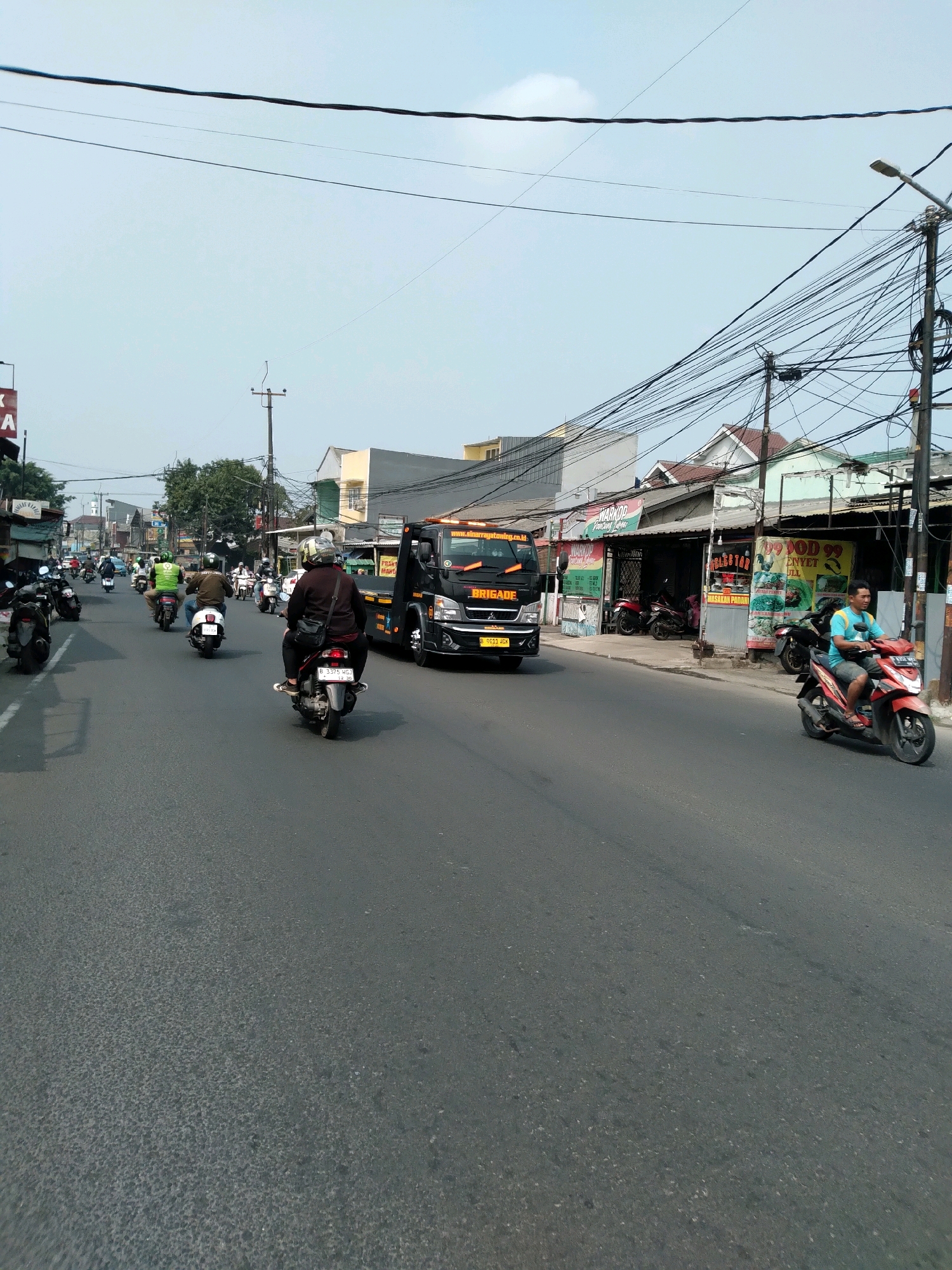 A wide, asphalt road in South Tangerang, Indonesia, is teeming with traffic, predominantly motorcycles and a tow truck. The sky is clear and pale blue. On the right side of the road, a row of small shops with colorful signs are visible, some with awnings extended over the sidewalk. Several parked motorcycles are in front of these establishments. A man in a blue t-shirt and dark shorts rides a red and black scooter, facing away from the camera and towards the right of the frame. Further down the street, a black tow truck with the word "BRIGADE" emblazoned on its side is moving towards the left, its headlights on. A license plate on the tow truck reads "B 0613 HGM". On the left side of the road, more motorcycles are traveling in the same direction as the tow truck. One rider, wearing a brown jacket and a white helmet, is on a white scooter. Another rider, in a green shirt and white helmet, is on a green motorcycle. Buildings line the left side of the street, interspersed with trees. Overhead, a dense network of electrical wires crisscrosses the scene, supported by poles. The road surface appears worn and has some texture.