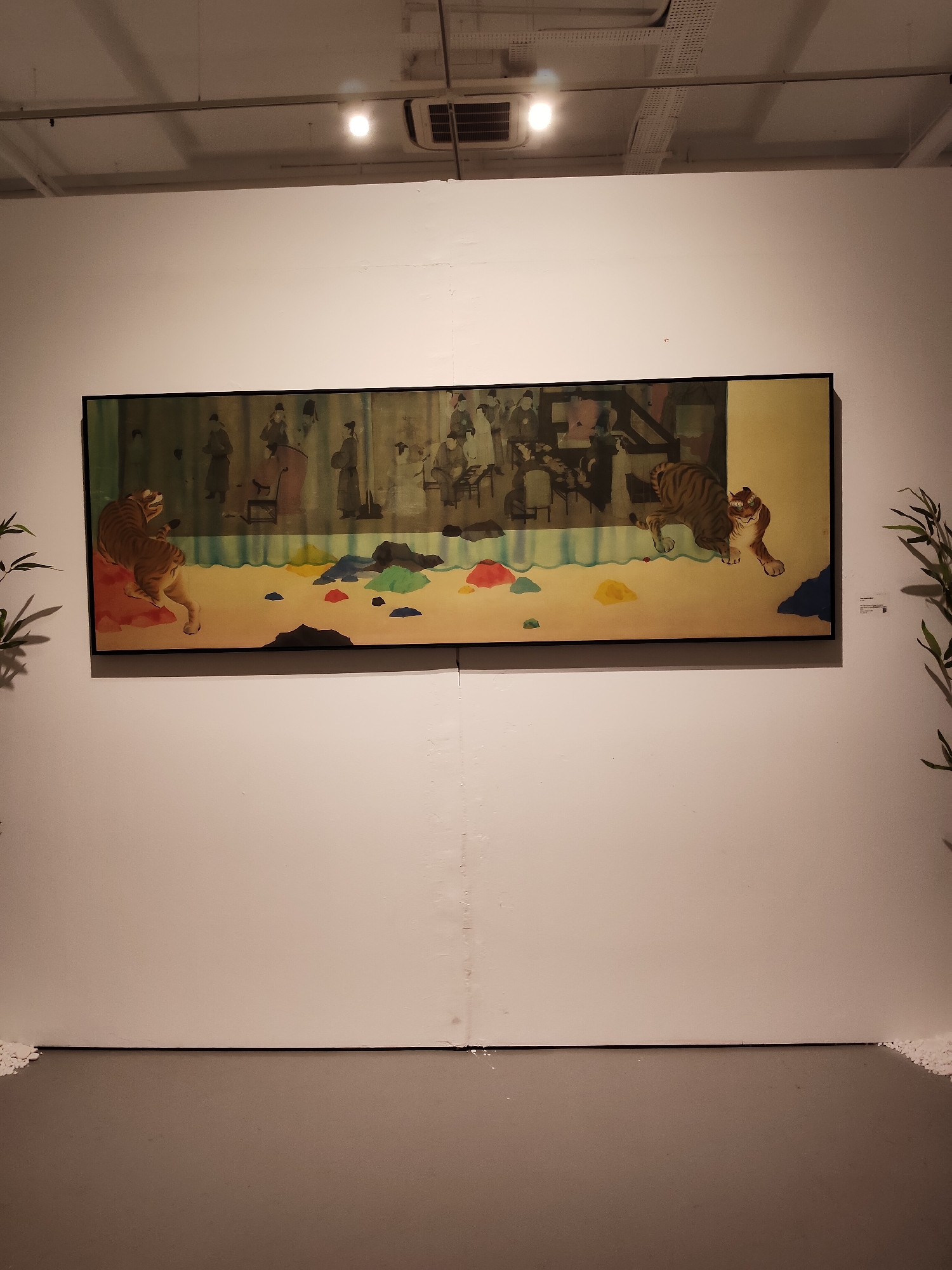 The image shows a painting hanging on a white wall in a gallery. The painting is a long horizontal artwork depicting a scene with tigers and people. On the left side of the painting, a tiger is seen sitting with its back to the viewer, looking towards the right. In the center and right side of the painting, there are several figures in traditional attire, possibly in a historical setting. Two tigers are prominently displayed in the foreground, one on the left and another on the right, both appearing to be in motion. The background of the painting is a muted green, while the foreground depicts a sandy or rocky terrain with scattered colorful shapes that could represent rocks or abstract elements. The painting is framed with a black border.
The gallery space appears to be modern, with track lighting on the ceiling and visible conduits. The wall is painted white and appears to have a horizontal seam running across it. In the foreground of the image, the floor is a dark gray. On the right side of the painting, a small white label is affixed to the wall, suggesting artwork information is present. In the far right and far left of the image, parts of green bamboo-like plants are visible, suggesting interior decoration or part of the exhibit. There is no visible information in the image to pinpoint the exact location within Singapore, Singapore, but it is clearly an art exhibition space.