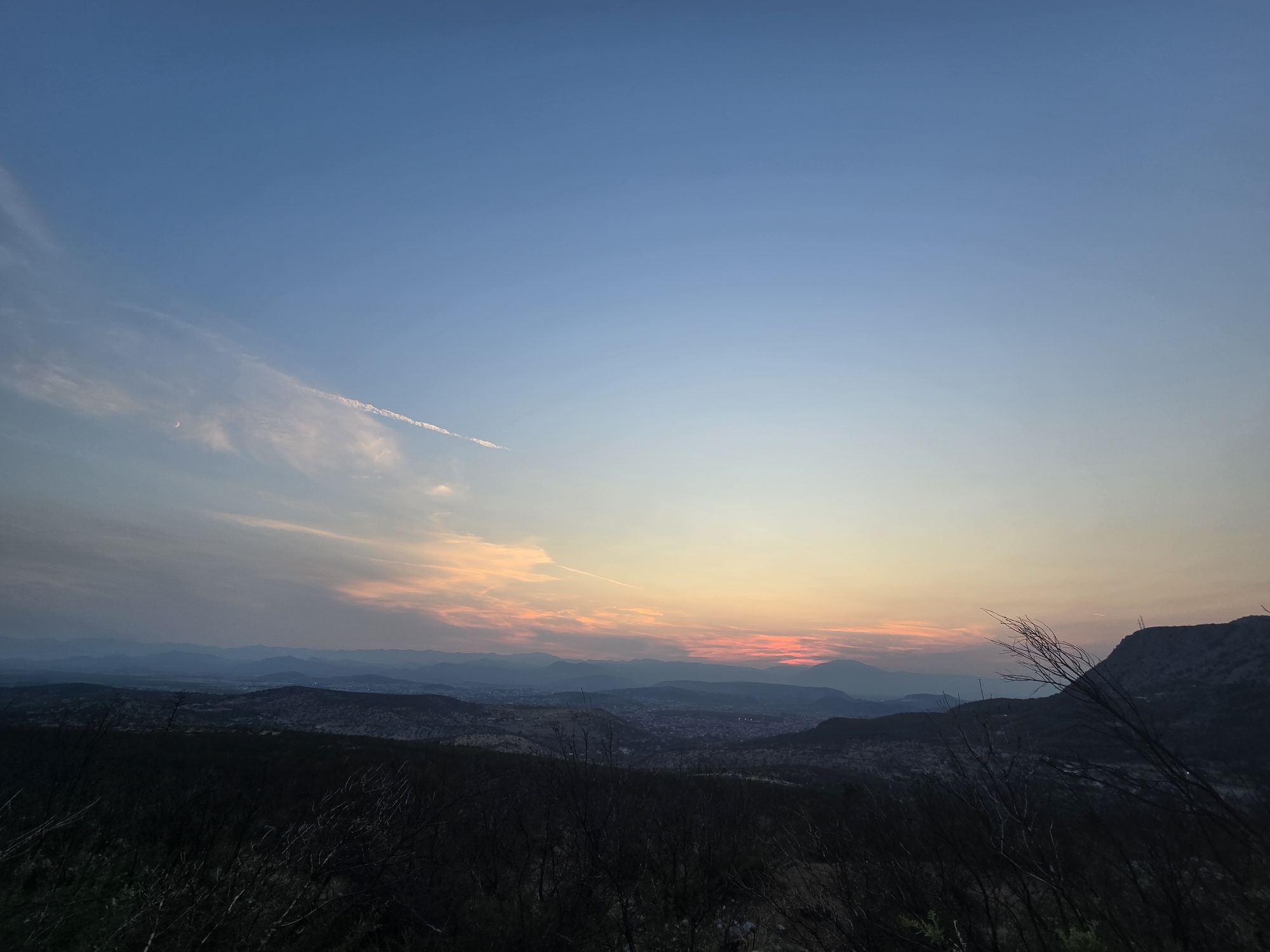 A wide-angle, horizontal landscape photograph captures a sunset over the city of Donja Vrbica, Montenegro. The sky transitions from a deep, clear blue at the top to lighter shades of blue, then to a band of orange and pink hues near the horizon, indicating the setting sun. Wispy clouds are scattered across the sky, with a faint contrail visible in the upper right portion. In the midground, layers of rolling hills and mountains recede into the distance, their shapes softened by atmospheric haze. The city of Donja Vrbica is visible nestled within the valley, appearing as a cluster of lights and structures. In the foreground, silhouetted against the illuminated sky, are dark, bare branches of trees, suggesting a late autumn or winter season. The overall scene evokes a sense of peaceful twilight and the vastness of the natural landscape surrounding the urban area.