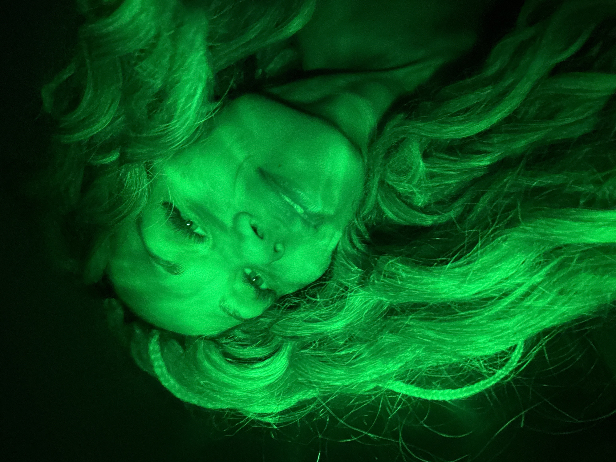 A green-toned, low-angle shot shows a person with light skin and long, wavy hair. The person's head is tilted, with their face angled towards the viewer. Their eyes are partially closed, and they appear to be smiling slightly. The background is dark, with the green lighting dominating the scene. The image appears to be artificially lit, resulting in the monochromatic green hue. The location in the city of Rusanovo, Russia, is not visually discernible from the image itself.