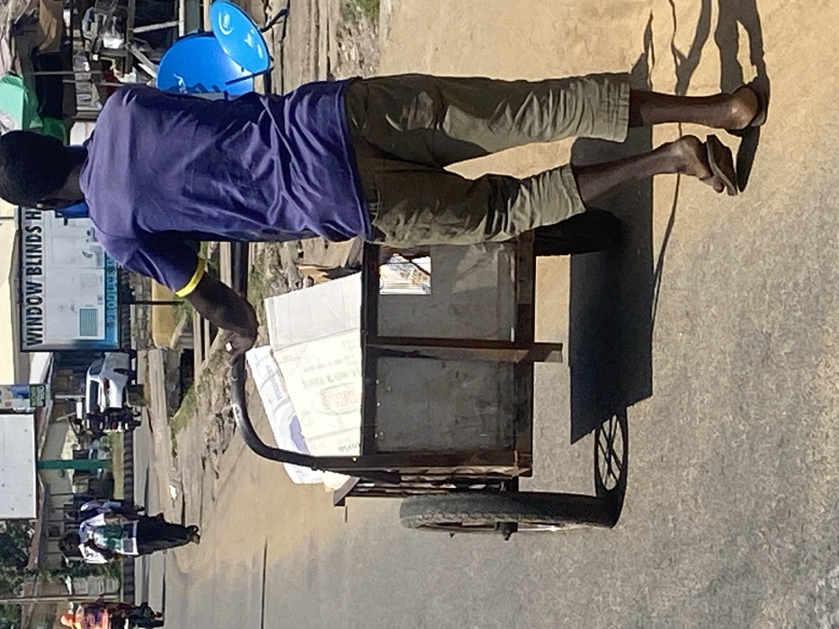 The image depicts a dark-complexioned male from behind, pushing a two-wheeled utility cart loaded with rectangular packaged goods on a paved street in Lokoja, Nigeria. The man wears a purple short-sleeved t-shirt with a yellow band on the right sleeve, olive green capri-length trousers, and brown flip-flops. The metal-framed cart contains multiple items, including boxes marked "YOGHURT" and "PRODUCED IN NIGERIA." In the midground-to-background, other pedestrians and motorcyclists are visible on the asphalt road to the left, along with a white car in the distance. To the right, a white commercial sign partially displays "WINDOW BLINDS" and "PRODUCE," positioned near two prominent blue satellite dishes and a green umbrella on the unpaved roadside. A small drainage channel runs parallel to the road. The scene is illuminated by bright daylight, casting clear shadows.