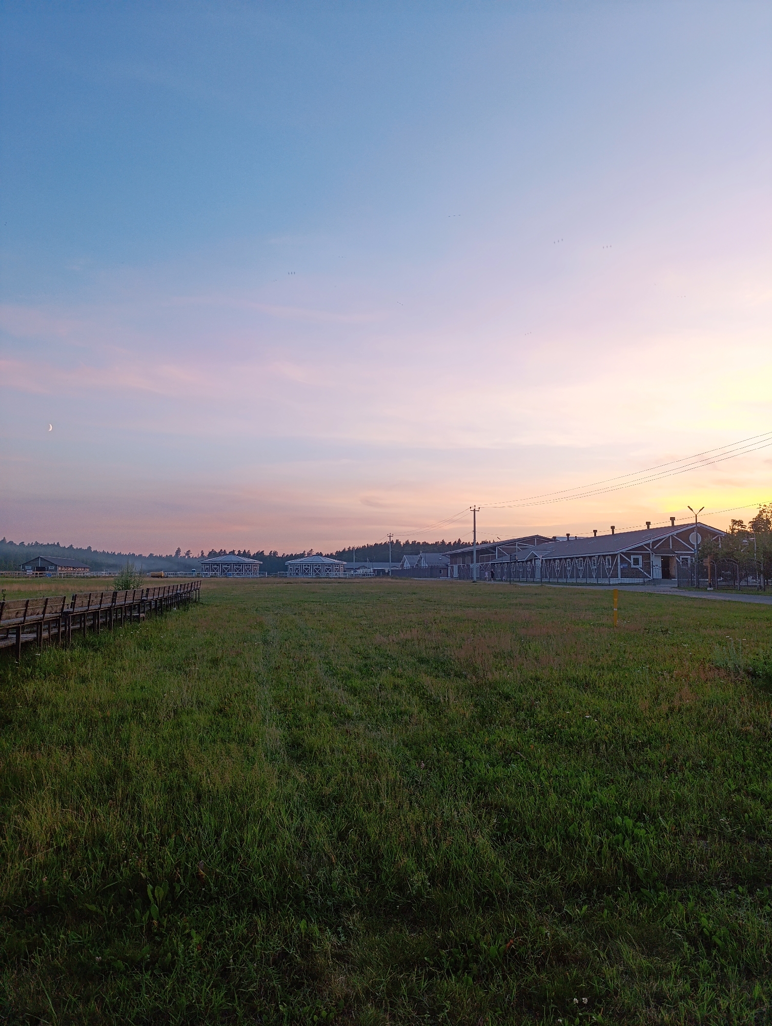 A wide-angle, eye-level shot captures a rural landscape at dawn or dusk. The sky is a gradient of soft blues and pinks, with a crescent moon visible in the upper left quadrant. In the middle ground, a long, low-lying building with a dark roof and white trim stretches across the frame. Several smaller, similar structures are visible to the left of the main building. A wooden boardwalk or fence runs along the left edge of the image, bordering a large, grassy field. The field is covered in lush green grass with some wildflowers. In the distance, a treeline marks the edge of the property, and utility poles and power lines are visible. Several small birds are silhouetted against the sky. The overall scene is serene and peaceful.