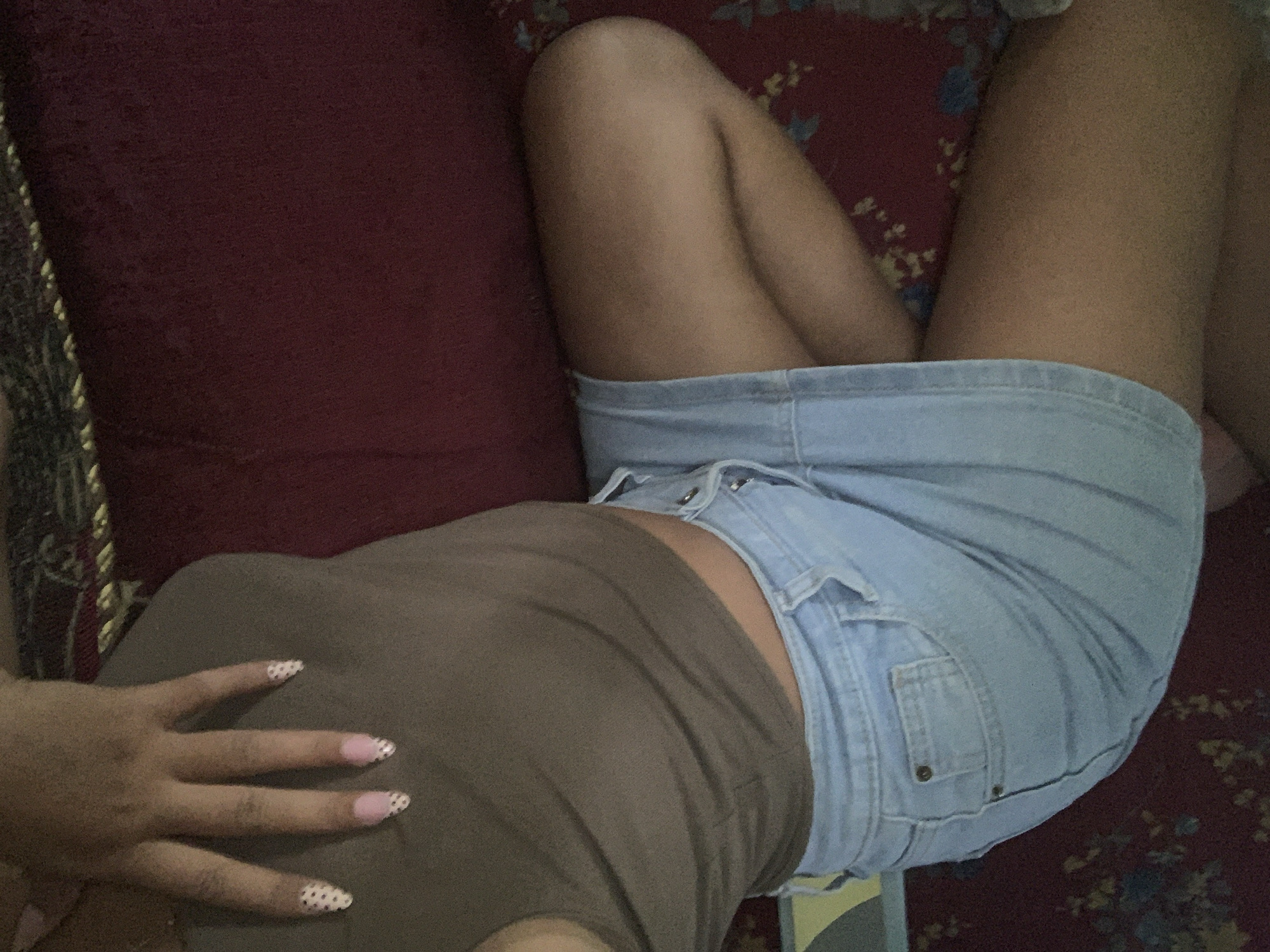 A close-up, overhead shot shows a person's torso and legs from the waist down, reclining on a red surface, possibly a couch or rug with a floral pattern. The person is wearing a brown, form-fitting tank top and a light blue denim skirt. The tank top appears sheer in some areas. The denim skirt has visible stitching and pockets. The person's left hand is visible in the upper left quadrant of the image, with manicured nails decorated with pink and white polka dots. The lighting in the image is dim. No specific location in Jajag, Indonesia can be determined from the visual information provided.