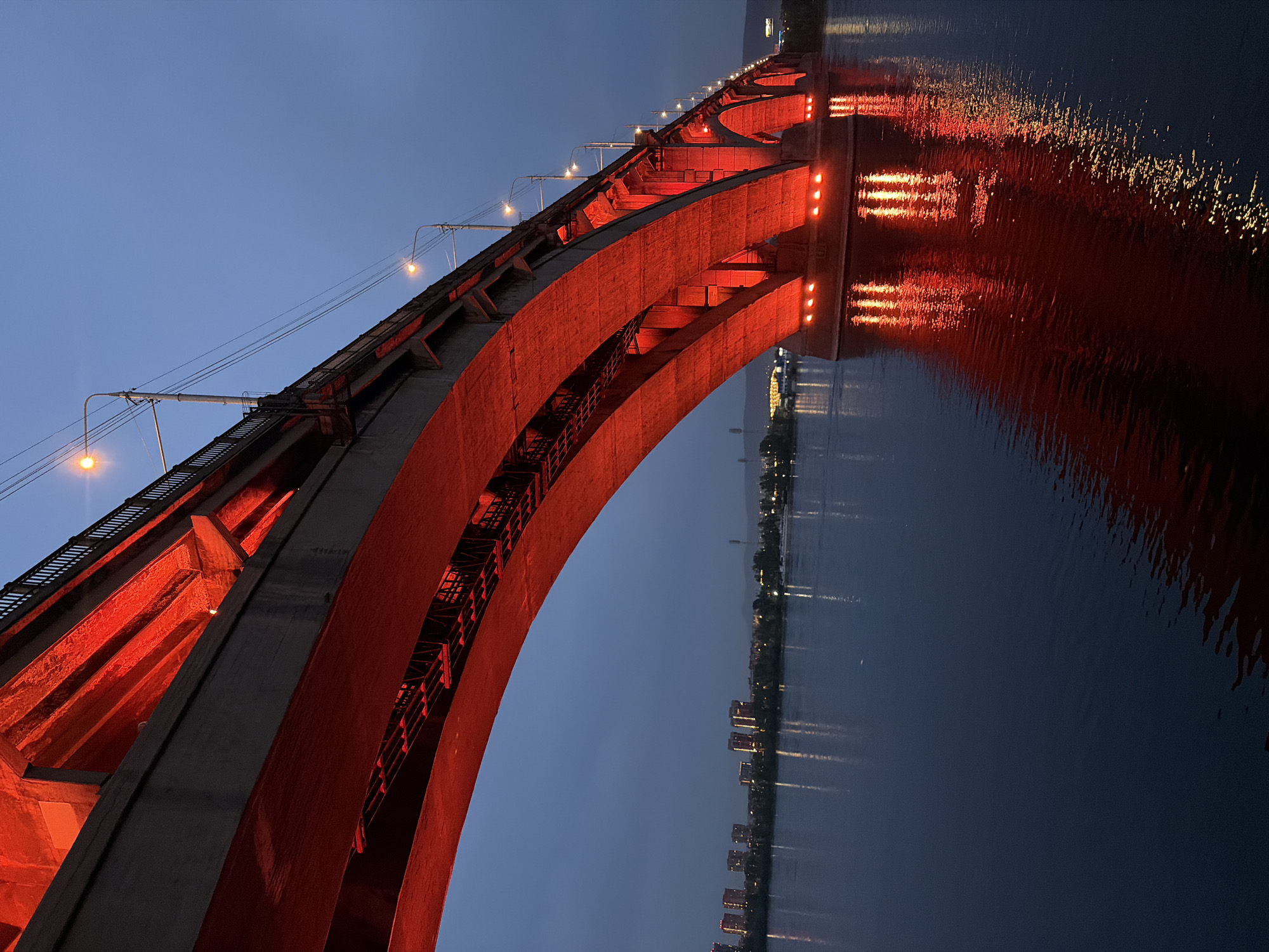 The image captures a wide view of the Krasnoyarsk Bridge, also known as the Yenisei Bridge, at dusk in Krasnoyarsk, Russia. The bridge, a massive concrete structure with a series of arches, is illuminated by vibrant red lights from below, casting a strong reflection on the dark, calm waters of the Yenisei River. The sky is a deep blue, transitioning from twilight. Along the distant riverbank, a cityscape with illuminated buildings and streetlights is visible, hinting at the urban environment. Several streetlights are also visible along the top of the bridge, casting their own yellow-orange glow. The overall scene is one of dramatic artificial lighting against a natural twilight backdrop, highlighting the architectural presence of the bridge over the river. There are no people or animals clearly visible in the image.