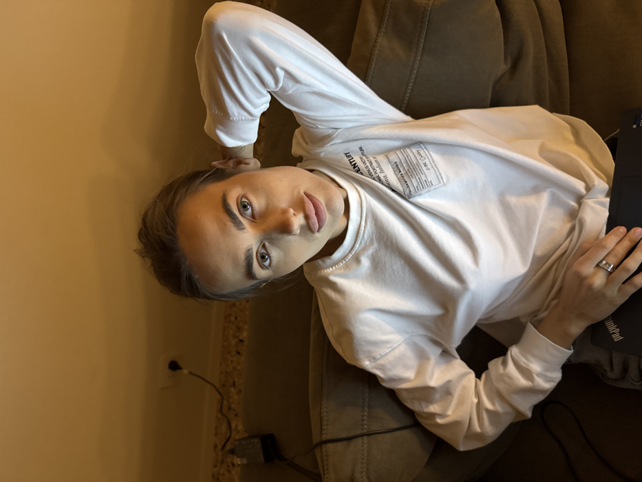 An indoor setting in Venice, Italy, depicts an adult female seated on a dark, upholstered sofa. She directly faces the camera. Her hair is styled away from her face, revealing prominent eyebrows, light-colored eyes, and full lips. She wears a white, long-sleeved crew-neck sweatshirt with a rectangular label-like patch on the front, left side. Text on the patch includes "CENTURY," "VENUS VICTOR," "ROMAN PORTRAIT CULTURE," "AURORA, ITALIANI," and "ITALIAN WRITER'S MANUSCRIPT." A silver-colored ring is visible on the fourth finger of her left hand, which rests on a black laptop, partially revealing a "ThinkPad" logo. Her right arm is raised, with the hand positioned behind her head/neck. The background features a plain, light-colored wall with a decorative trim below it. In the upper left, a white electrical outlet is visible, with a black power cord plugged in and extending downwards to a dark power adapter resting on the sofa. Warm, artificial light illuminates the scene from above the subject.