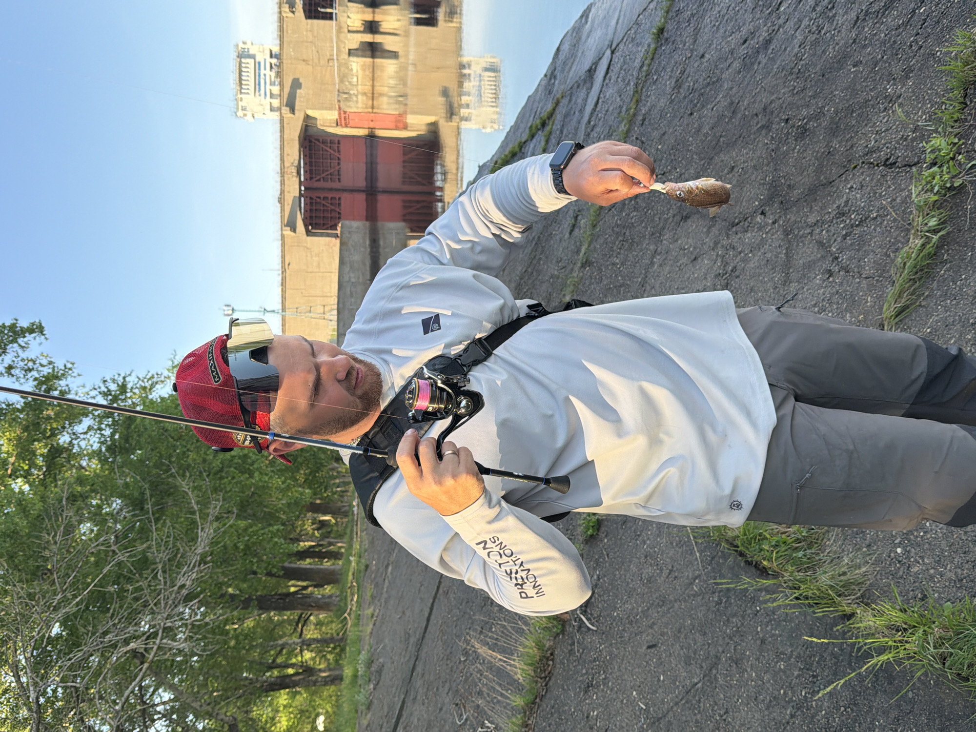 A man is fishing at the Votkinsk Dam in the city of Chaikovsky, Russia. He is wearing a light gray long-sleeved shirt, gray pants, a red hat, and sunglasses. He holds a small fish in one hand and a fishing rod in the other. The fishing rod has a pink reel. A strap is across his chest. He is standing on a concrete surface near patches of grass. The Votkinsk Dam with its red gates is visible in the background, along with its reflection in the water. Trees line the left side of the image. The sky is light blue.
