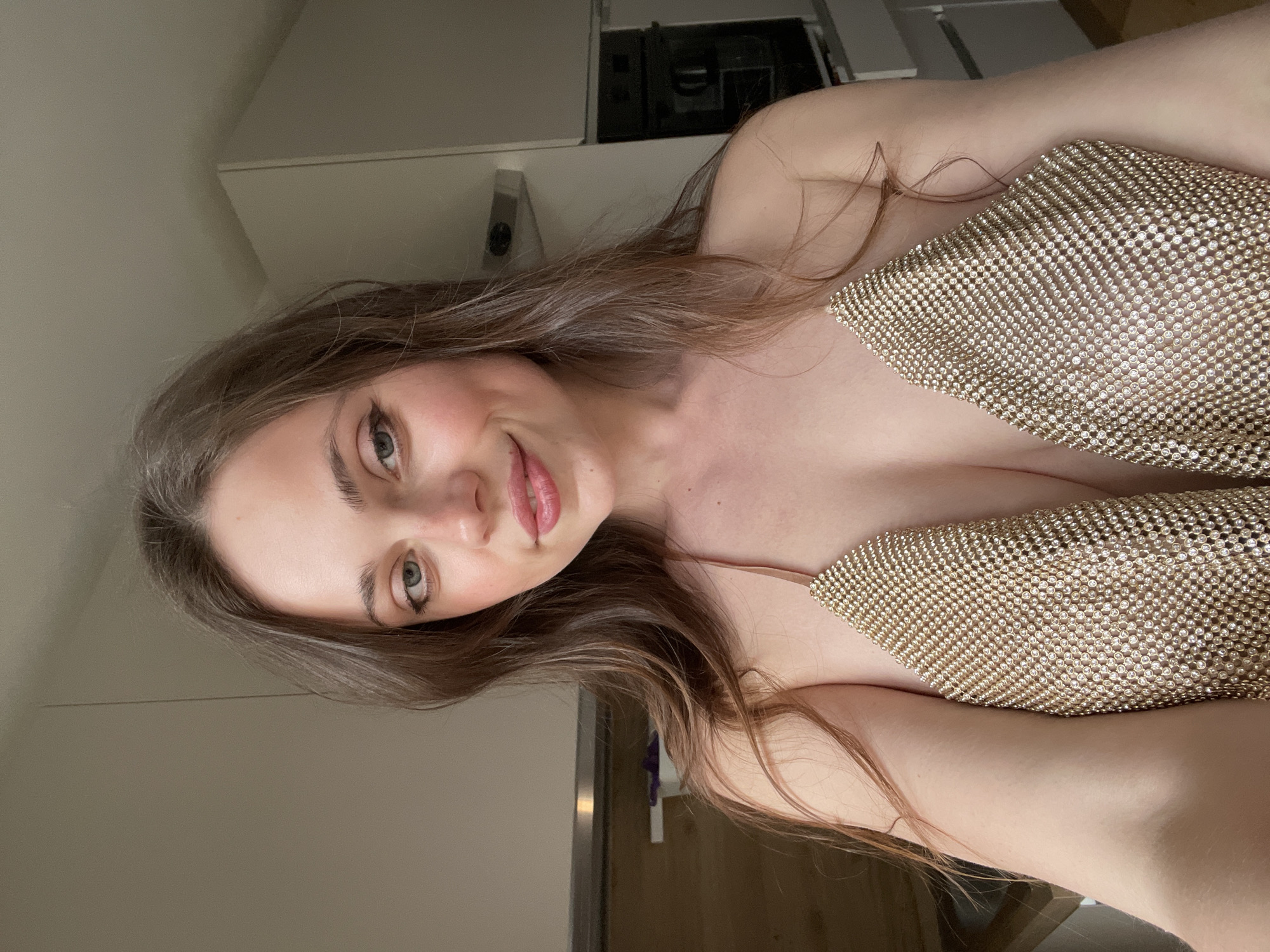 A young woman is posing for a selfie indoors. She has long brown hair and is wearing a sparkling gold top. The background shows elements of a modern kitchen, including cabinets and appliances. The location cannot be determined to be Sappada, Italy, based on the given image content.