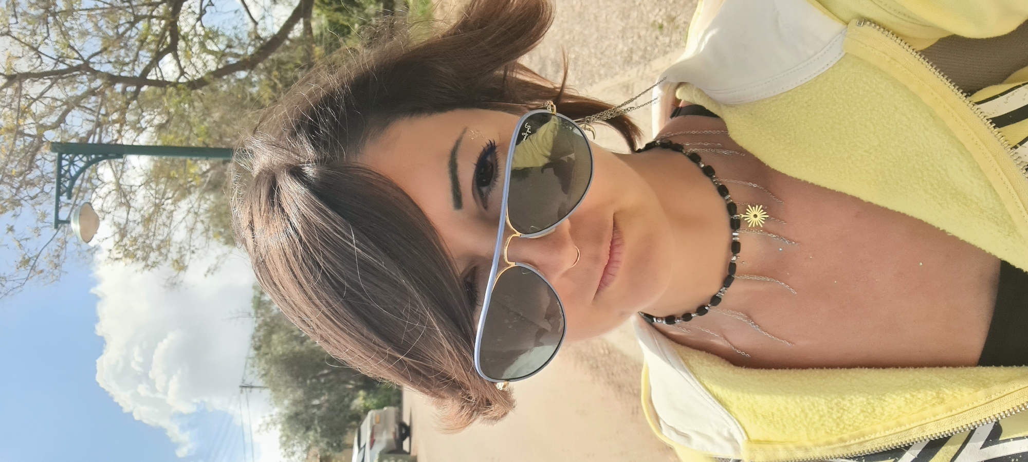 A woman is posing outdoors in Lachish Regional Council, Israel. She is wearing sunglasses, a necklace, and a yellow jacket. The background includes trees, the sky with clouds, and street lighting.