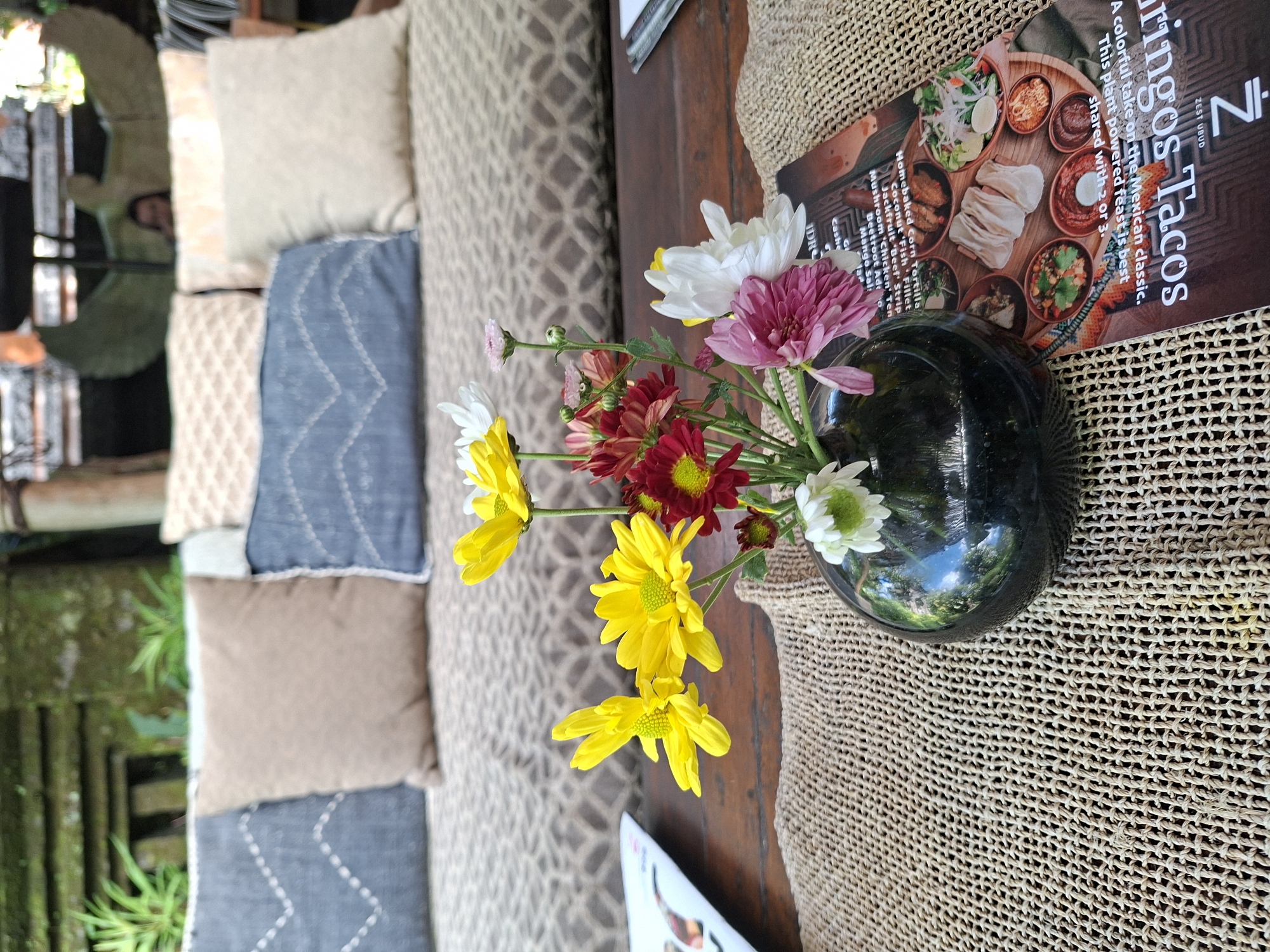 The image presents a serene and inviting outdoor or semi-outdoor dining and lounge area, characteristic of a cafe or restaurant in Ubud District, Indonesia, under natural daylight.
In the foreground, a dark wooden table is partly covered by a beige woven mat. A small, glossy black vase holds a vibrant bouquet of chrysanthemums, featuring bright yellow, white, pink, and red/maroon blossoms. Adjacent to the vase is a menu or flyer titled "Zest Ubud Gringos Tacos," which states: "A colorful take on the Mexican classic. This plant powered feast is best shared with 2 or 3." The menu also lists various plant-based ingredients and dishes, including "Homebaked Corn Tortilla," "Coconut 'Fish Fillet'," "Jackfruit 'Beef' Strip," "om 'chic ken Ten," "Beetroot Ade," "Mango Sal," "Cashe," and "DR".
To the left, a comfortable bench seat is adorned with several cushions of varying patterns and solid colors, including beige, blue with white stitching, and brown, suggesting a cozy and relaxed seating arrangement. The background is lush with green foliage and what appears to be bamboo, reinforcing the tropical location. No people are clearly visible in the scene. The overall ambiance is calm and welcoming, highlighting a tranquil space for dining or relaxation.