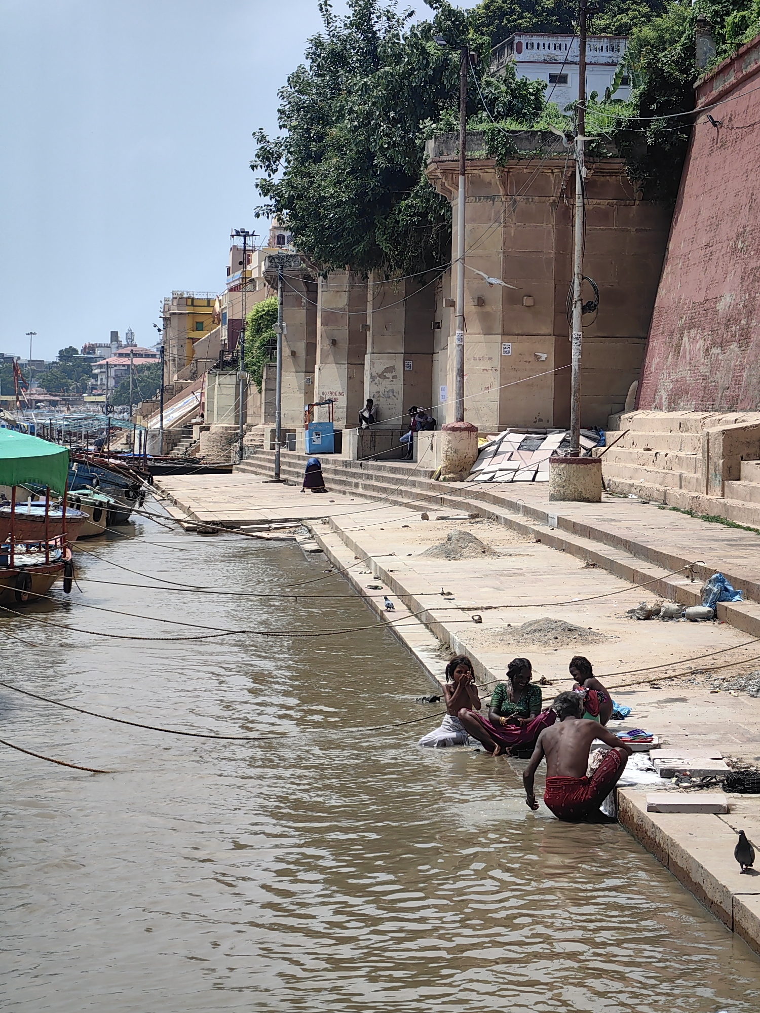 The image captures a scene along the ghats of Varanasi, India, a city renowned for its sacred riverbanks. In the foreground, a muddy, brown river flows gently. Along the bank, a concrete walkway is visible, with several people present. Three young individuals, two girls and a boy, are sitting in the water at the edge of the ghat. The boy is in the process of washing himself, wearing red patterned trousers and a red cloth around his waist. The girls are also in the water, one appearing to be speaking with the others, and another covering her mouth. Another individual, a woman or older girl, is seated on the ghat's edge, also in red clothing.
Further up the ghat, there are several boats moored to the bank, some with green awnings. Beyond the boats, a series of stone steps leads up to buildings that line the river. These buildings are of various colors and architectural styles, with some featuring intricate stonework and balconies. Trees with lush green foliage are interspersed among the structures, and the sky is a clear blue. The overall impression is of a bustling, daily life unfolding by the holy river.