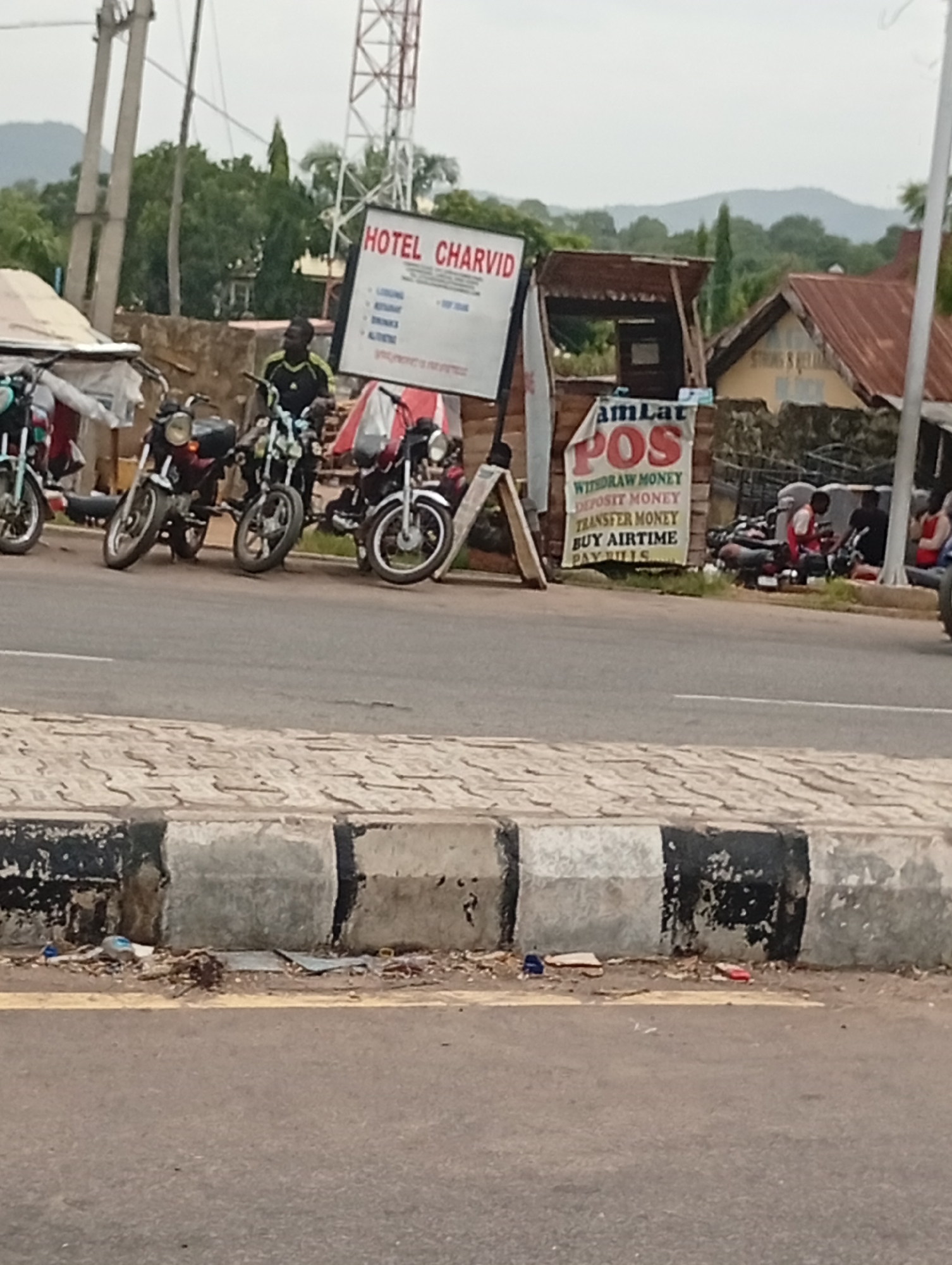 The image displays a roadside scene in Adankolo, Nigeria, under an overcast sky. In the foreground, a paved road runs parallel to a concrete curb, sections of which are painted black and white. Litter is scattered on the ground adjacent to the curb.
The middle ground features a row of several motorcycles, parked in a line, suggesting a commercial motorbike stand. A man wearing a black and green shirt stands next to one of these motorcycles, looking towards the right. Further to the right and slightly behind, two other individuals are visible near more parked motorcycles, one in a red top and another in a dark top.
Two signs are prominent. A large white sign, mounted on a wooden A-frame, advertises "HOTEL CHARVID" and lists services such as "LODGING", "RESTAURANT", "DRINKS", and "ALTITUNES" (likely amenities or activities). To its right, a banner-style sign displays "amLat POS", detailing financial services available: "WITHDRAW MONEY", "DEPOSIT MONEY", "TRANSFER MONEY", "BUY AIRTIME", and "PAY BILLS". This indicates a local point-of-sale service center.
In the background, various buildings with reddish-brown corrugated iron roofs are visible amidst abundant green trees and vegetation. A tall telecommunications mast or tower stands on the left side, reaching into the sky. Distant hills or mountains line the horizon. The scene depicts a lively, semi-urban commercial area, likely during the daytime given the ambient light, though without direct sunlight.