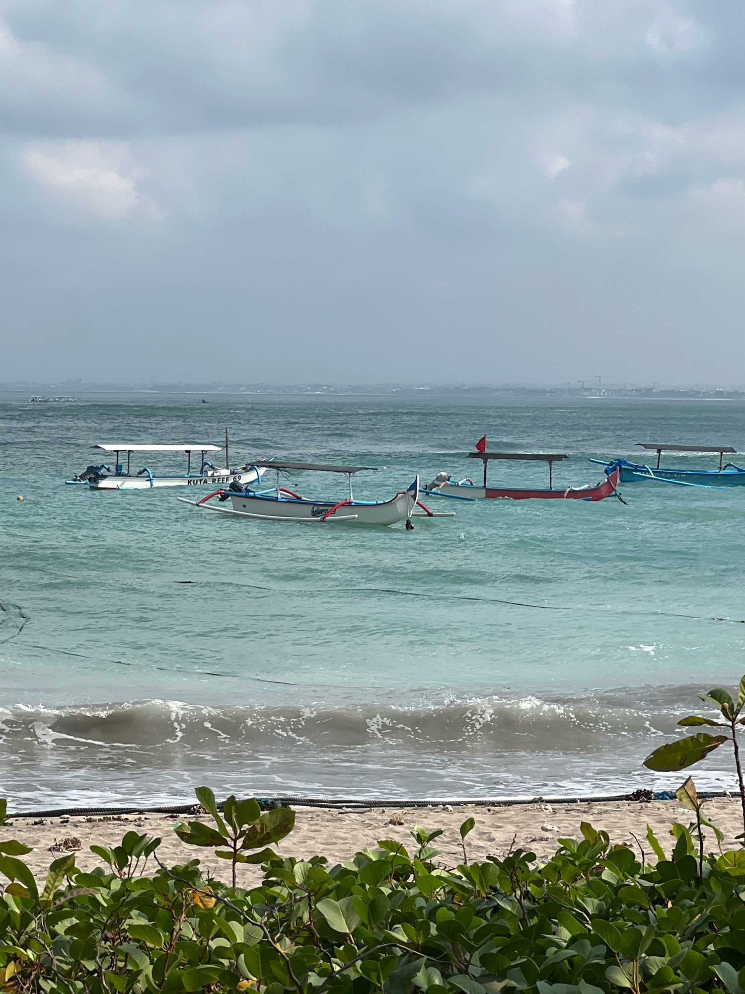 This image shows a view of the ocean from the beach in Kuta, Indonesia. Several traditional Indonesian outrigger boats are anchored in the turquoise water. The boats have wooden hulls and are equipped with canopies for shade. One of the boats in the foreground has the text "KUTA REEF 69" visible on its side. In the distance, a hazy coastline is visible, with some structures suggesting an urban area. The sea is relatively calm with gentle waves approaching the sandy beach. Lush green foliage occupies the foreground, framing the scene. The sky is overcast with light clouds.
