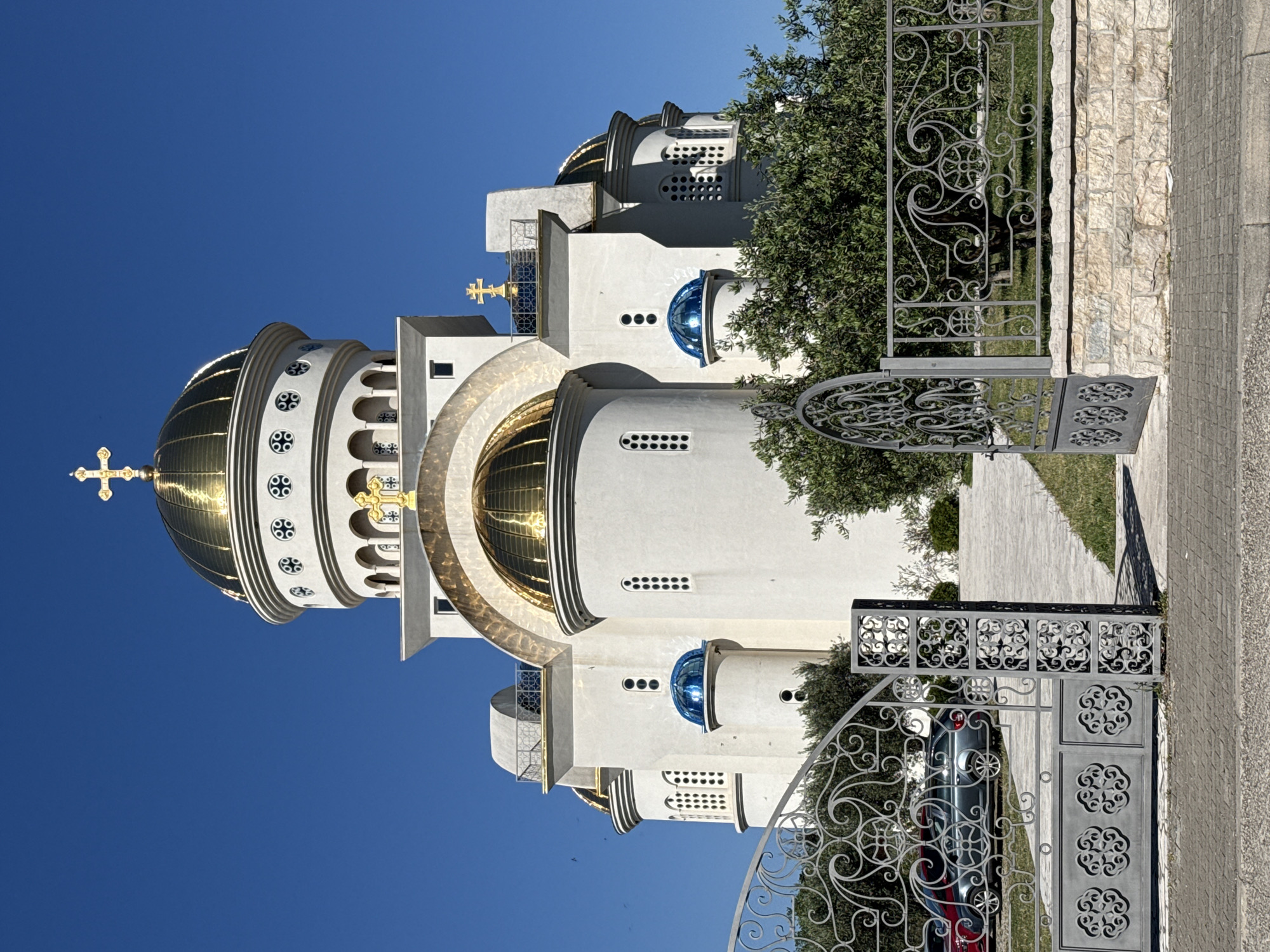 The image shows the St. John Vladimir's Church in Bar, Montenegro, viewed from behind an ornate metal gate. The church is a white building with golden domes, adorned with golden crosses. One visible dome is large and prominent. The building also has arched windows and other architectural details. A tree grows near the church, partially obscuring a section of the building. The sky is a clear, bright blue. The gate is partially open, allowing a glimpse of a car parked on the other side. A sidewalk is visible in the foreground.