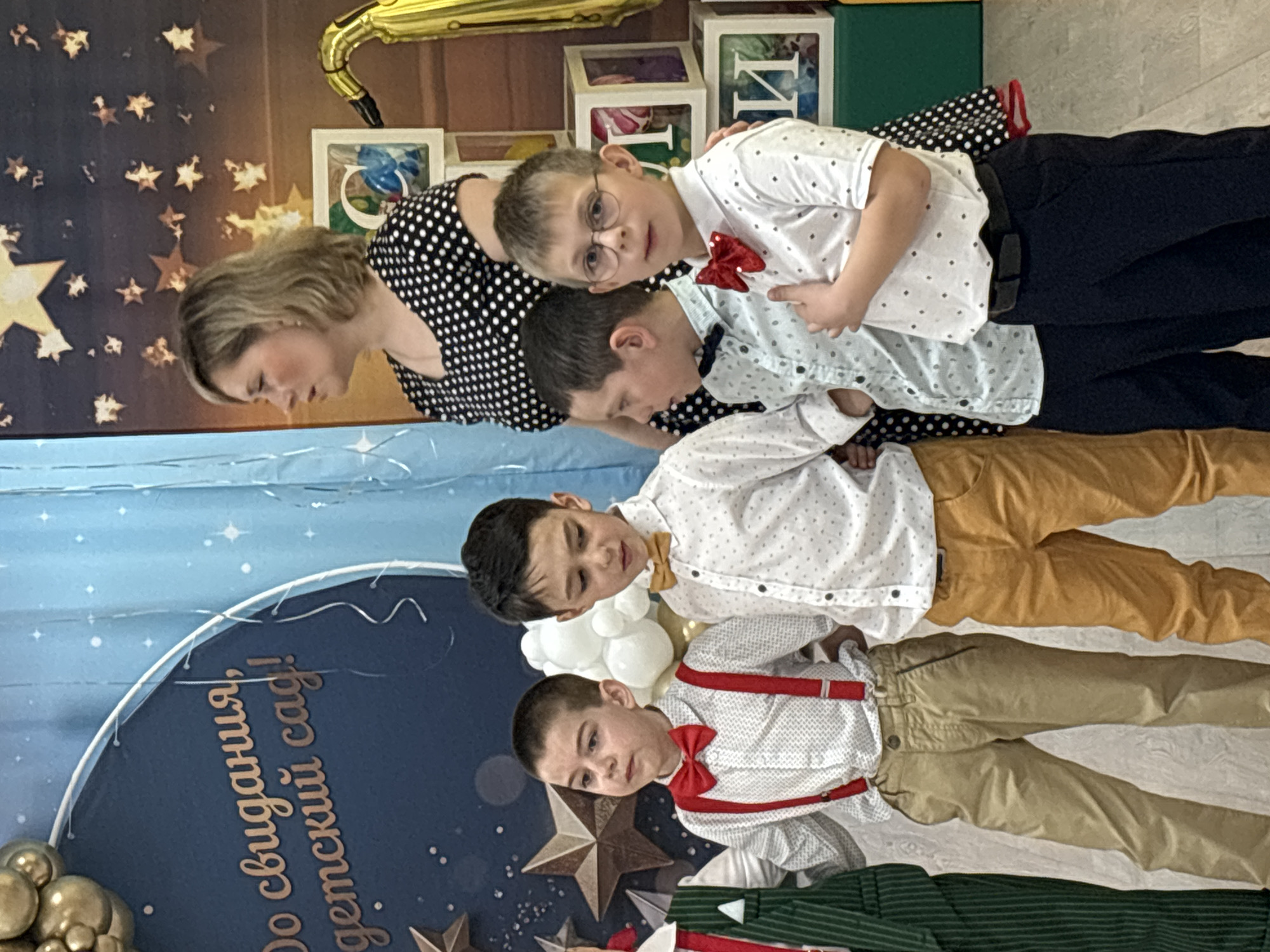 A woman and four young boys are posing for a picture in the city of Unknown, Unknown. The woman is wearing a black and white polka dot dress. The boys are all wearing white shirts and various colored pants. One boy is wearing glasses and a red bow tie. Another boy is wearing a blue bow tie. One boy is wearing a yellow bow tie and the fourth boy has a red bow tie with red suspenders. They are standing in front of a blue banner with the words "До свидания, детский сад!" in gold lettering, which translates to "Goodbye, kindergarten!". There are stars and balloons visible in the background.