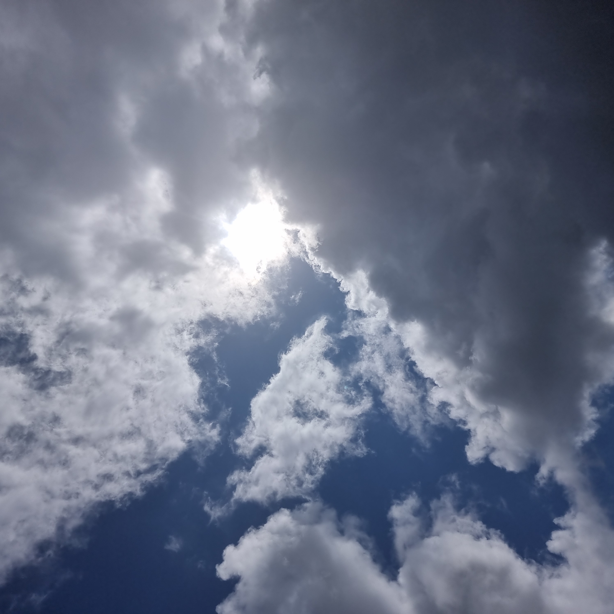This is a photograph of a dynamic sky, likely taken in Dunnae-myeon, South Korea, as suggested by the location context. The image is dominated by a mixture of dark, ominous storm clouds and brighter, fluffy cumulus clouds, interspersed with patches of clear blue sky. The sun is partially obscured by a thick bank of clouds, creating a dramatic and somewhat diffused light. This suggests that the time of day is likely midday or afternoon, when the sun would be at its highest point. The presence of both dark and light clouds indicates variable weather conditions, possibly transitioning from or towards a storm. There are no people or specific objects visible, making the sky itself the sole subject. The overall mood conveyed by the image is one of dramatic natural beauty and the power of the elements. The texture of the clouds varies from smooth and wispy to heavy and turbulent. There is no visible text in the image.