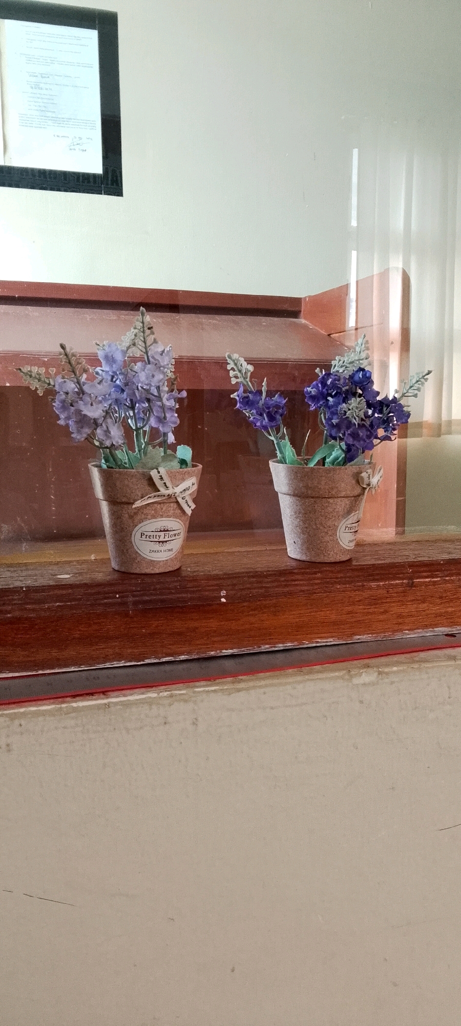 The image depicts two small decorative potted plants with artificial flowers, likely lavender or similar, placed on a wooden shelf. The pots are light brown and appear to be made of a cork-like material, each adorned with a small ribbon and a label. The left pot has a label that reads "Pretty Flower ZAKKA HOME" and the right pot has a similar label. The flowers are in shades of purple and blue, with some greyish-green foliage. Behind the plants, there is a dark wooden background that appears to be part of a shelf or display cabinet. The shelf itself is also made of dark wood and has a reddish-brown trim along the front edge. To the left, a framed document is partially visible, hanging on a light-colored wall. The overall environment suggests an indoor setting, possibly a home or a shop, and the lighting indicates it is daytime. The location is identified as Unknown, Indonesia.
