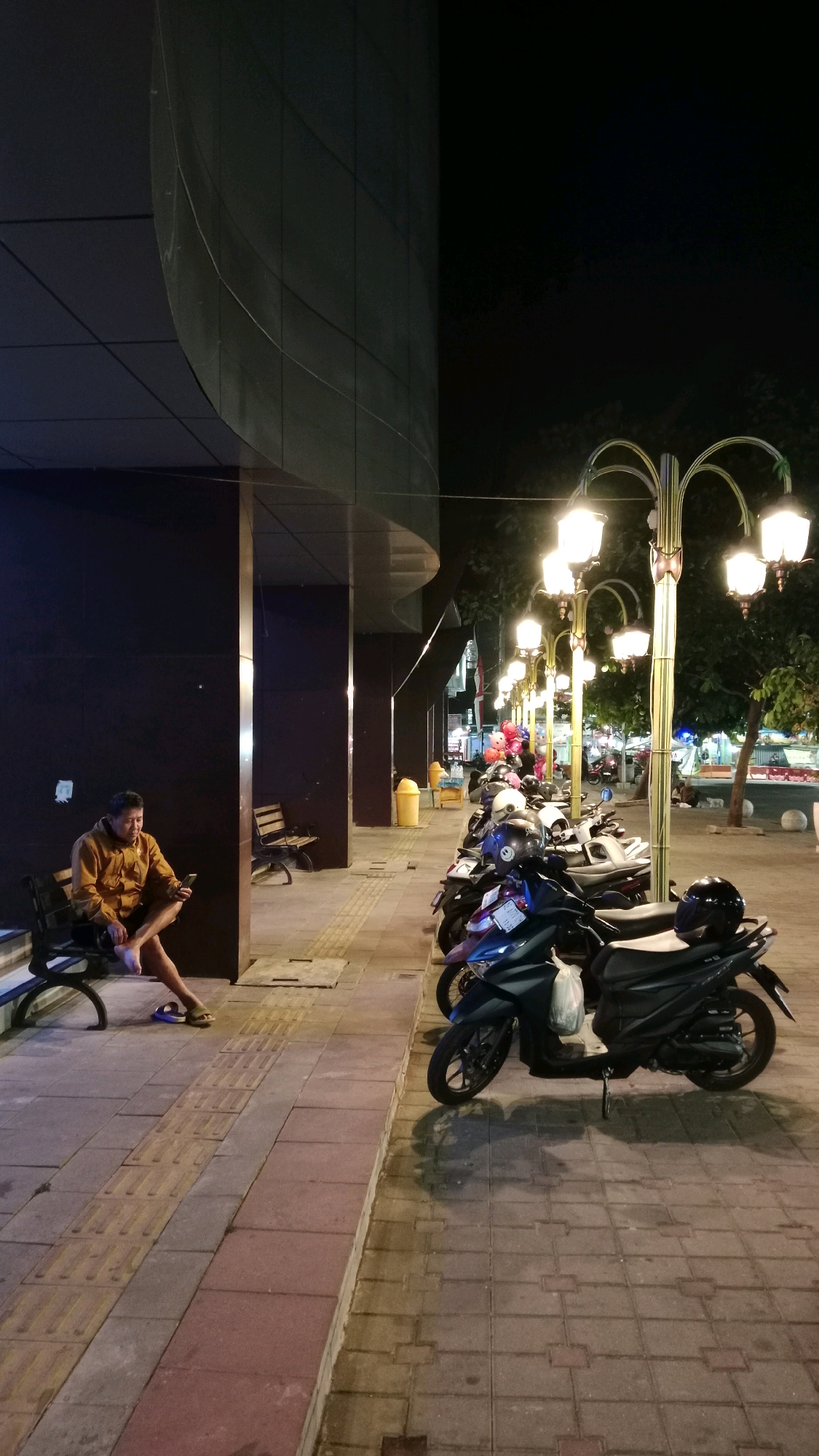 A man is sitting on a bench outside a building at night, looking at his phone. He is wearing a mustard-colored jacket and dark pants. He is barefoot and wearing sandals. In front of him, a row of motorcycles are parked along a sidewalk. Streetlights illuminate the scene, casting shadows on the ground. The building behind him has a dark, curved facade. In the background, there are more lights and trees. The area appears to be a public street or plaza in Magetan, Indonesia.
