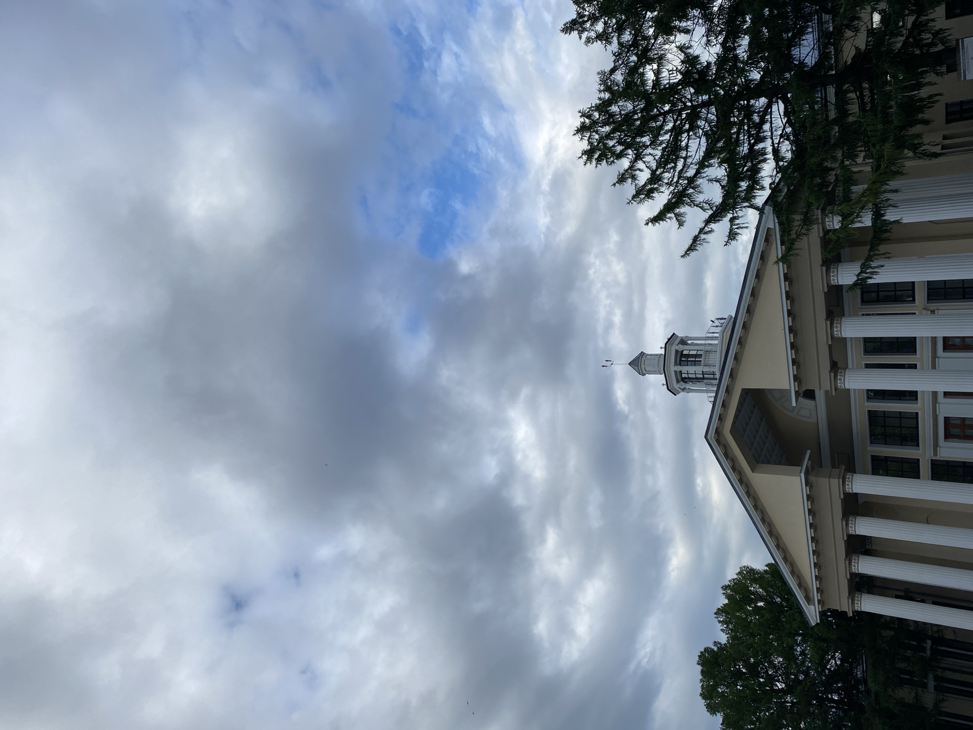 This media file captures an upward-looking view of a building with classical architectural elements, set against a cloudy sky. The building features white columns and a light-colored facade, with dark-framed windows visible. A portion of a dome or tower structure is present at the top of the building, topped with what appears to be a flag or weather vane. Overhanging branches of a dark green tree are visible in the foreground and on the right side of the frame, partially obscuring the building and the sky. The sky is filled with a mix of gray and white clouds, with some patches of blue sky peeking through, suggesting overcast or partly cloudy weather. The overall lighting suggests it could be daytime, likely late afternoon or early morning given the cloud cover. The orientation of the image is portrait. There are no people or discernible activities taking place. The scene evokes a sense of grandeur and possibly a slightly somber or dramatic atmosphere due to the cloudy sky. No text is visible in the image. Based on the provided location context of Kazan, Russia, this building could be a historical landmark, government building, or public institution.