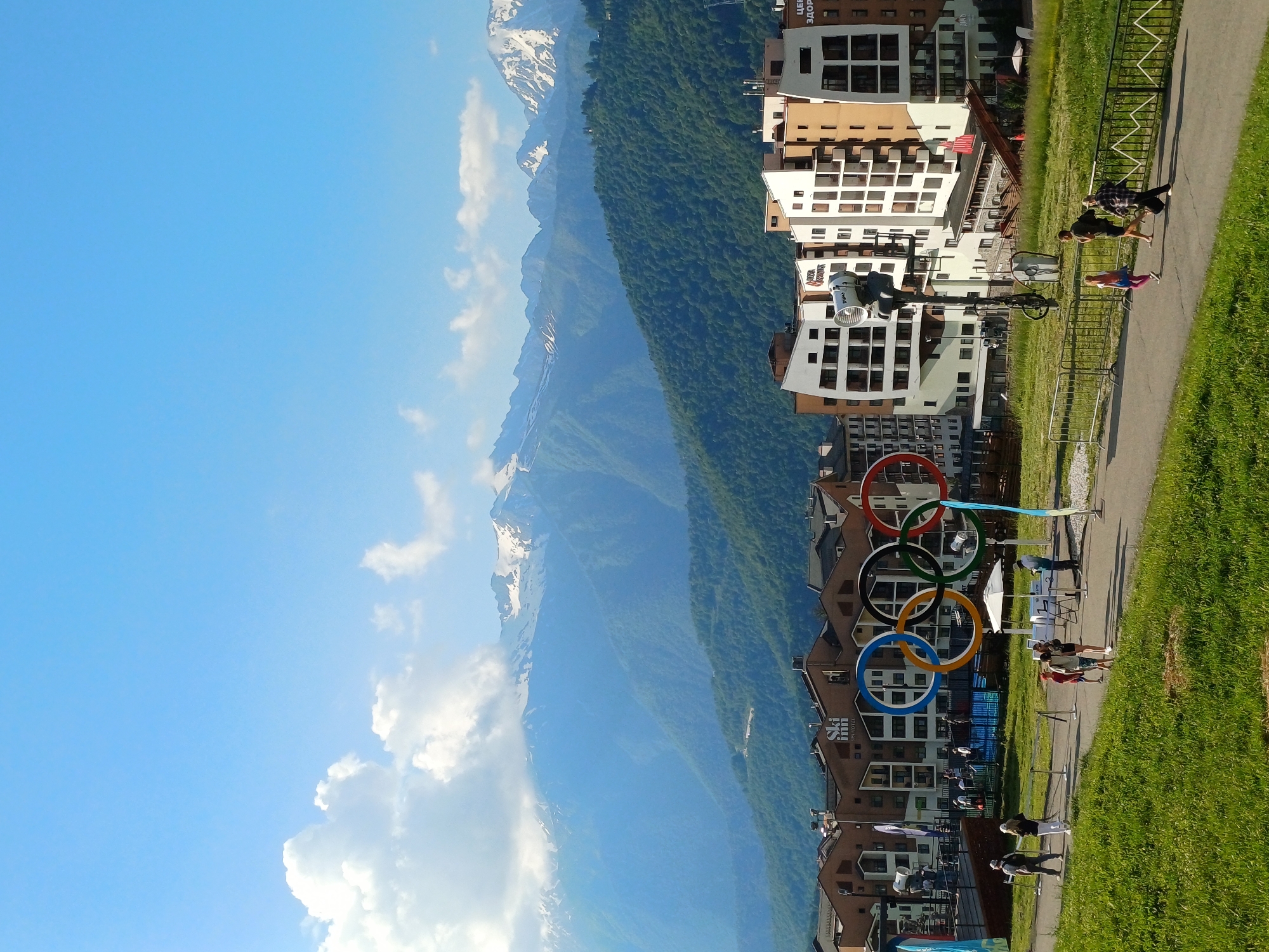 This image captures a scenic view of a mountain resort with several people engaged in outdoor activities. The scene is set against a backdrop of majestic, snow-capped mountains and lush green slopes. Multiple buildings, likely hotels or residences, are visible, characterized by their modern architecture with balconies and white and brown facades.
Prominently displayed in the foreground is a large Olympic rings sculpture, a clear indicator of the location being related to Olympic events or a legacy site. Several individuals are scattered around, some walking along a paved path adjacent to a grassy area, while others appear to be exercising or practicing yoga. One person is seen jogging, another stretching, and a group of people are gathered near the Olympic rings.
The sky is a brilliant blue with scattered white clouds, suggesting a pleasant, sunny day. The time of day appears to be late afternoon or early evening, given the quality of the light and the shadows cast. The overall atmosphere seems relaxed and active, with people enjoying the beautiful natural surroundings and taking advantage of the recreational opportunities offered by the resort.
Visible text includes "Ski Inn" on one of the buildings and some Cyrillic text that appears to be "Центр здоровья" (Center of Health) on another. The combination of Olympic symbolism, mountain scenery, and resort architecture strongly suggests this is a location associated with winter sports and tourism, specifically in Esto-Sadok, Russia, which hosted events during the 2014 Sochi Winter Olympics.
