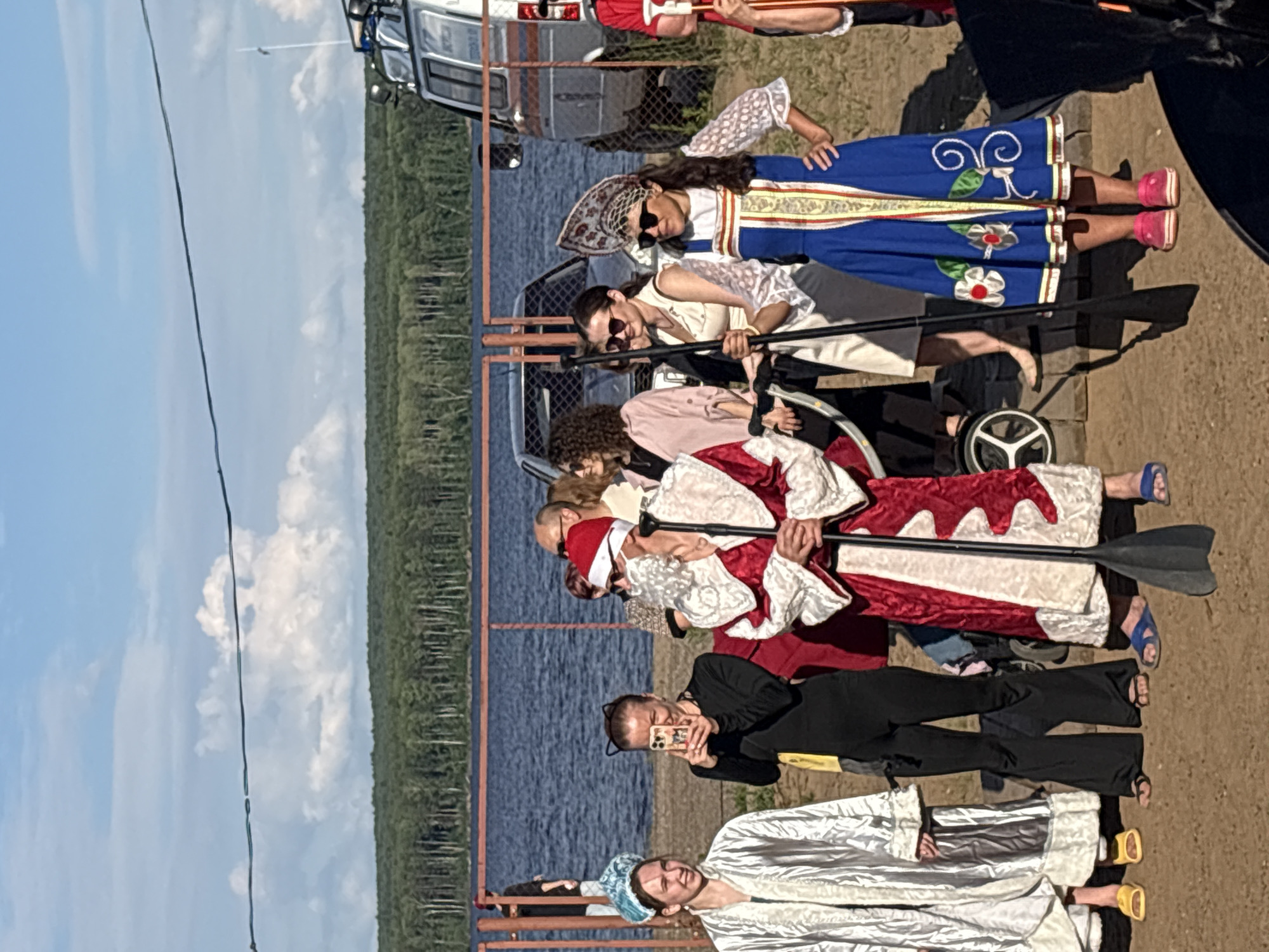 A group of people are gathered outdoors near a body of water. In the foreground, on the left, a woman in a silver costume and blue headwear stands. To her right, a woman in a black outfit with cat ears is holding up a smartphone, appearing to take a photo. Next to her, a person dressed as Santa Claus, complete with a red and white costume and beard, holds a paddle. To the right of Santa, a woman in a blue and white traditional Russian dress and headdress is also present. Behind them, other individuals are visible, along with a fence, vehicles, and trees in the background. The scene appears to be set on a sunny day, likely in the city of Chaikovsky, Russia, given the context of the traditional attire and the presence of water.