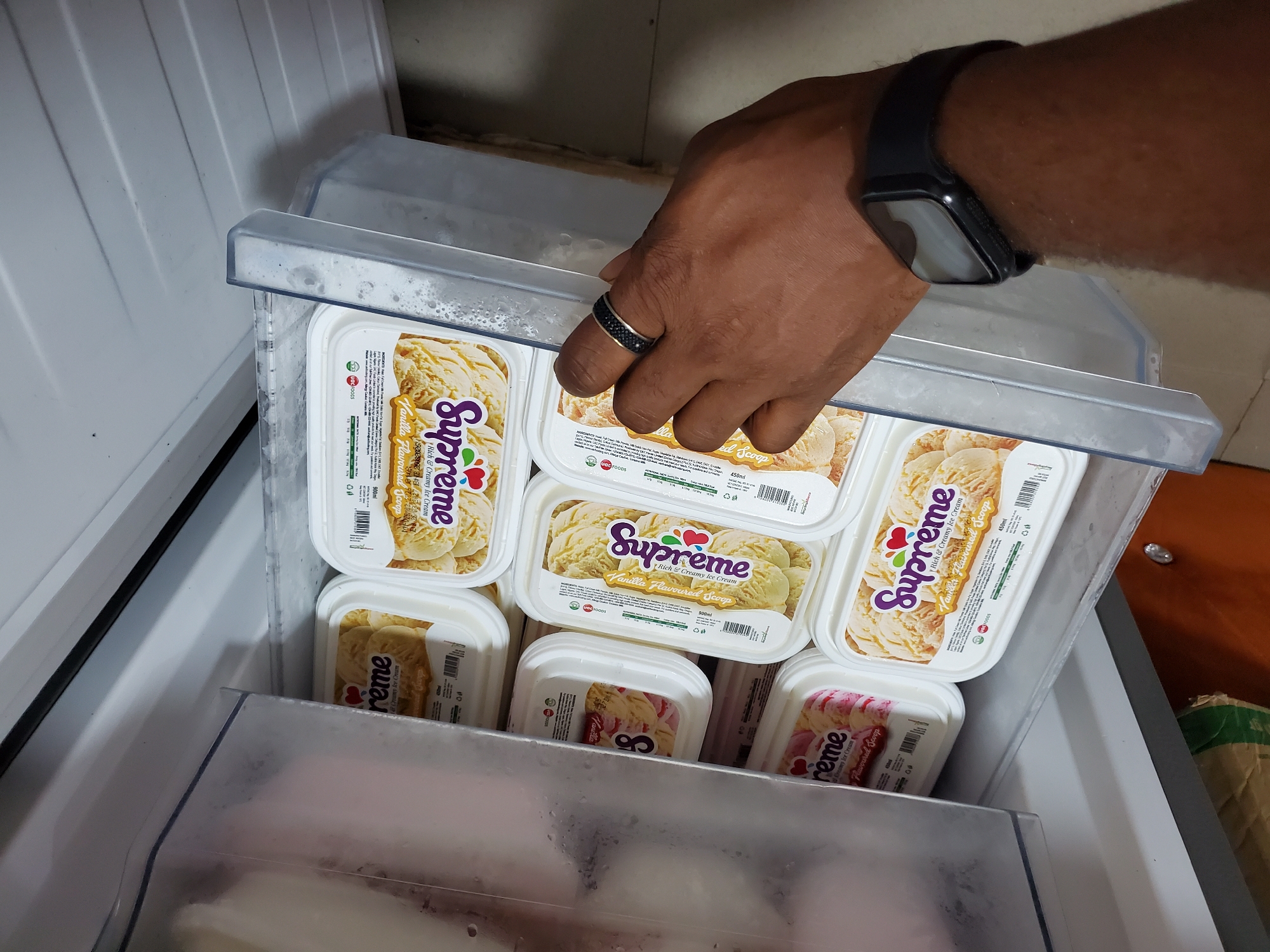 The image captures a close-up, high-angle view inside an opened freezer, likely located in Nkpor, Nigeria. A person's right arm and hand, characterized by dark skin, extend into the freezer's interior, holding up a clear, frosted plastic lid or tray. A black ring is visible on the person's ring finger, and a black smartwatch is worn on the wrist.
The freezer is densely packed with multiple rectangular plastic tubs of ice cream. The majority of the tubs are "Supreme" brand, prominently displaying "Rich & Creamy Ice Cream" and "Vanilla Flavoured Scoop." These vanilla tubs are a light yellow-cream color and also feature "UGG FOODS" as the manufacturer/distributor, with some indicating volumes like "450ml" or "900ml." Beneath these, other ice cream tubs are partially visible, including some with pink and white swirls, suggesting a strawberry or mixed berry flavor, also under the "Supreme" brand. A smaller, partially obscured tub also shows "Vanilla" and the number "8."
The activity depicts the person interacting with the freezer, possibly retrieving, storing, or organizing the ice cream. The setting appears to be an indoor environment, given the artificial lighting, consistent with a kitchen or a retail establishment. No specific time of day or emotions are discernible.
