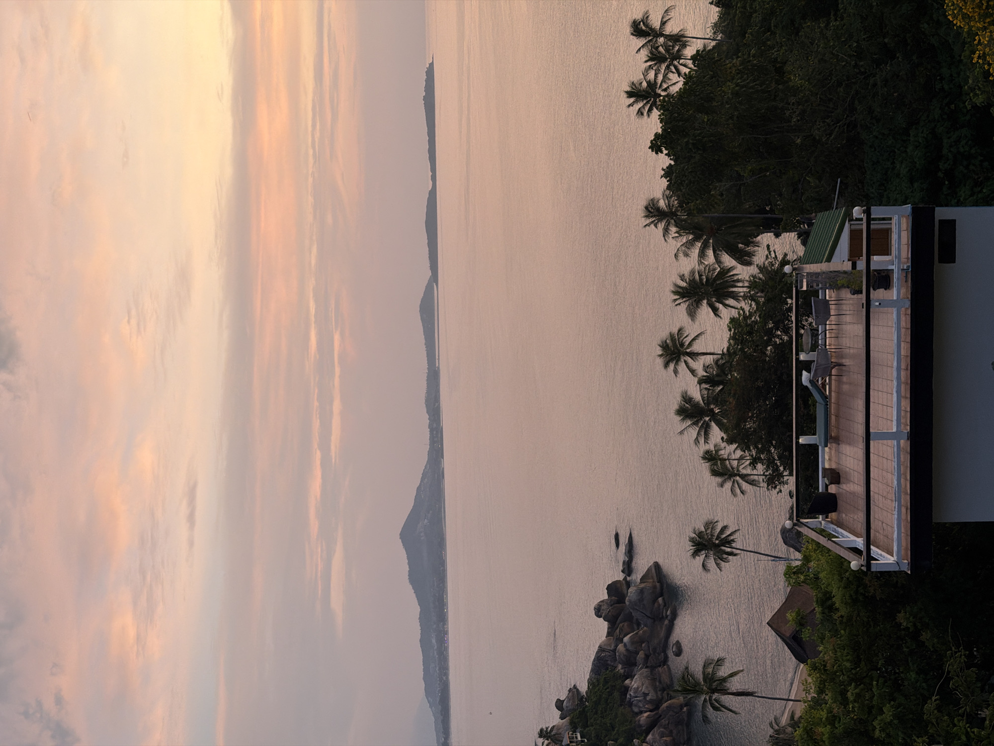 The photograph depicts a scenic overlook at sunset in the Ko Samui District of Thailand. The scene is dominated by a serene ocean stretching towards a distant mountain range. In the foreground, a modern rooftop terrace with white railings and tiled flooring offers a vantage point for the view. The terrace is furnished with a lounge chair and a table with chairs, suggesting a place for relaxation. Lush green vegetation, including palm trees, frames the terrace and slopes down towards a rocky coastline with boulders. A small, dark-roofed building is partially visible nestled amongst the trees below the terrace. The sky above is painted with soft hues of pink and orange, characteristic of a tropical sunset.