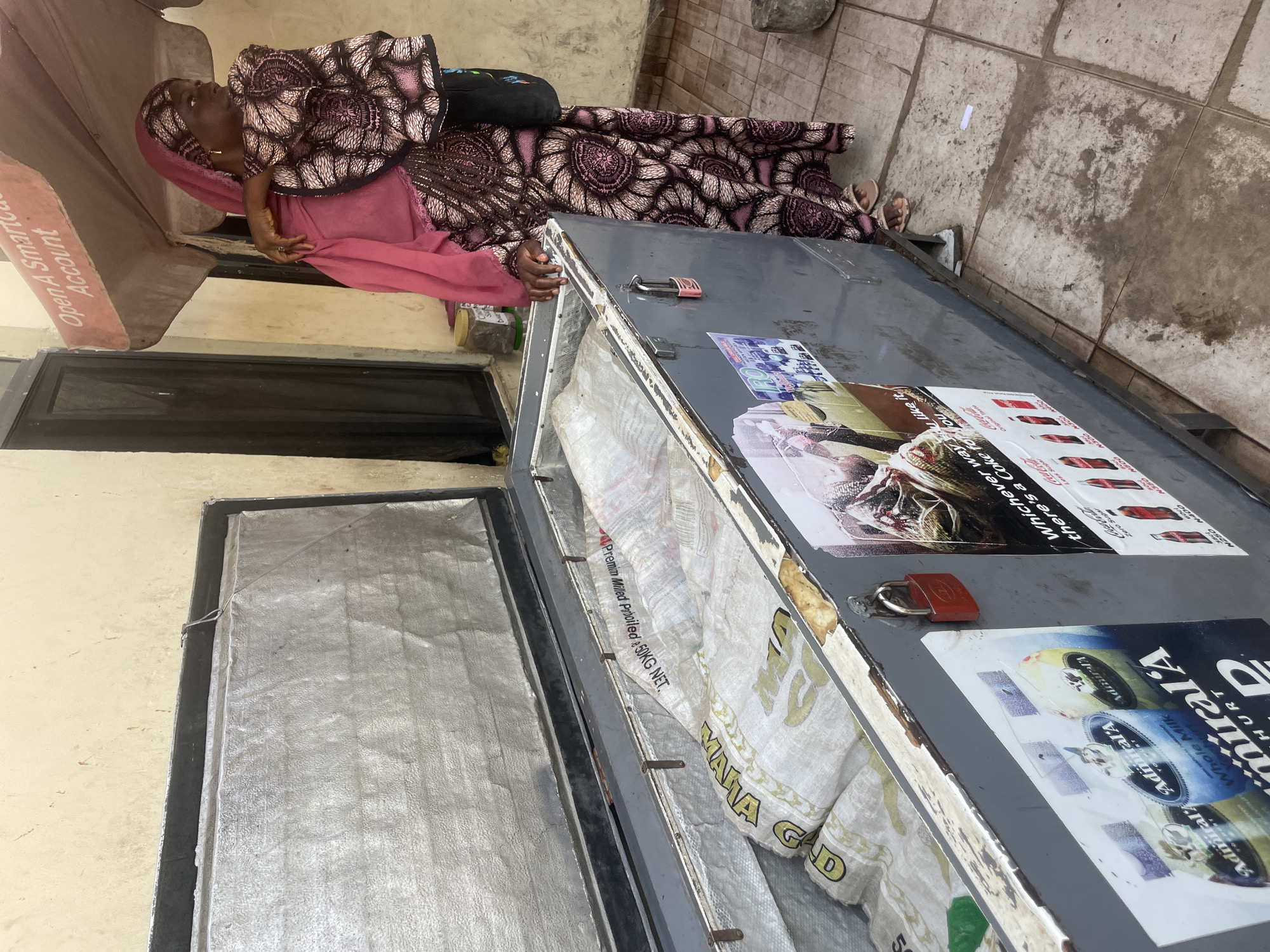 A woman in a pink headwrap and a patterned dress stands next to an open, gray chest freezer filled with bagged goods. The freezer is on a tiled outdoor area, and behind it, a wall and a window are visible. A portion of a red umbrella with the words "Open A SmartCard Account" printed on it is visible above the woman. The woman is looking away from the camera, her right arm resting on the edge of the freezer. The freezer has several stickers on its side, including one for Coca-Cola and a poster featuring a bird. There are also two padlocks attached to the freezer. The environment appears to be a marketplace or street vendor stall in Bauchi, Nigeria.