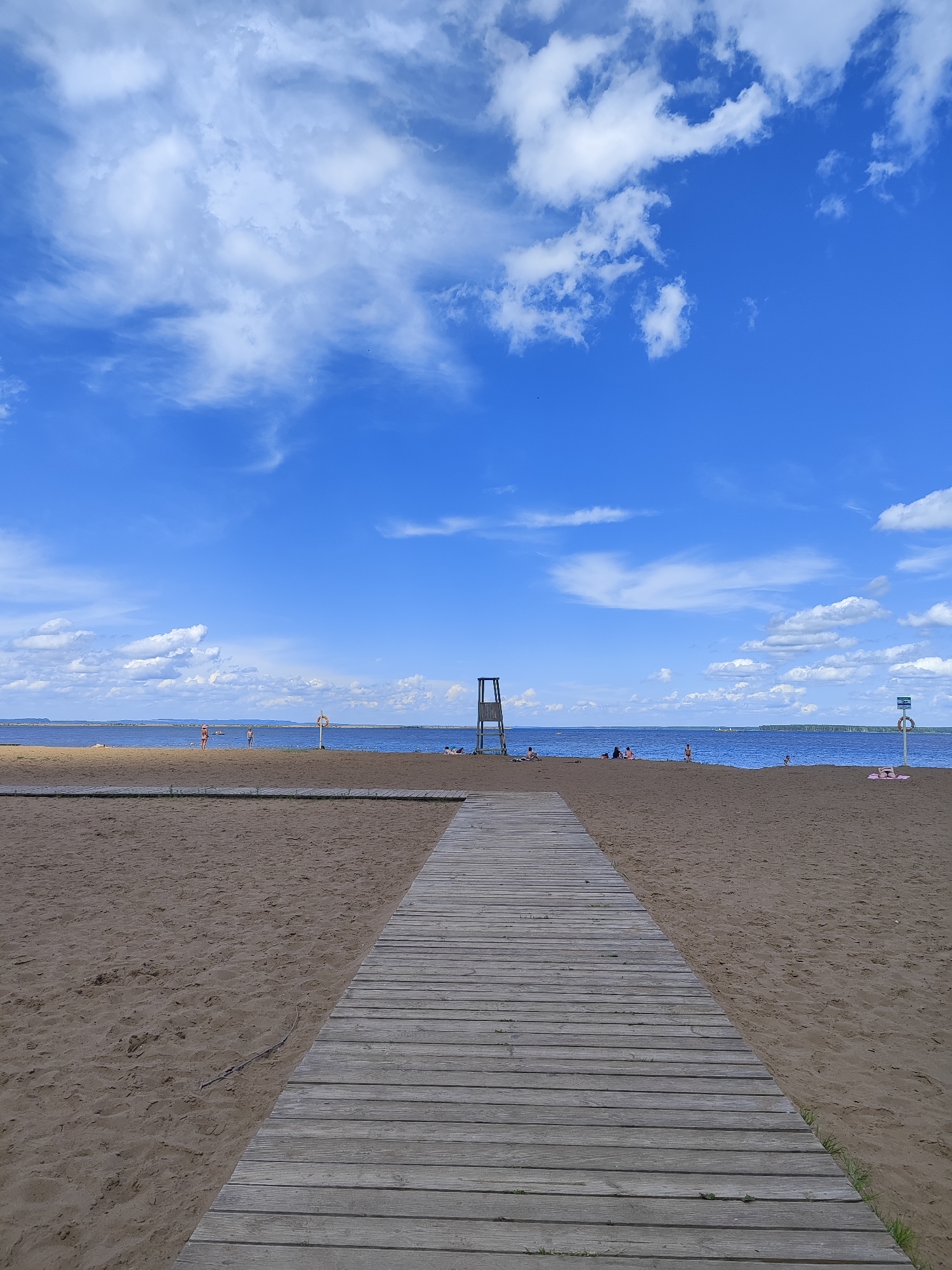 The image depicts a sandy beach with a wooden boardwalk leading towards the water under a bright blue sky with scattered white clouds. Several people are visible on the beach and in the water, some appearing to be sunbathing or swimming. A lifeguard tower stands on the right side of the beach, and a life buoy is mounted on a pole nearby. In the distance, land can be seen across the water. The scene is consistent with a public beach location, likely within the city of Chaikovsky, Russia, given the context of the provided hints which indicate this setting.
