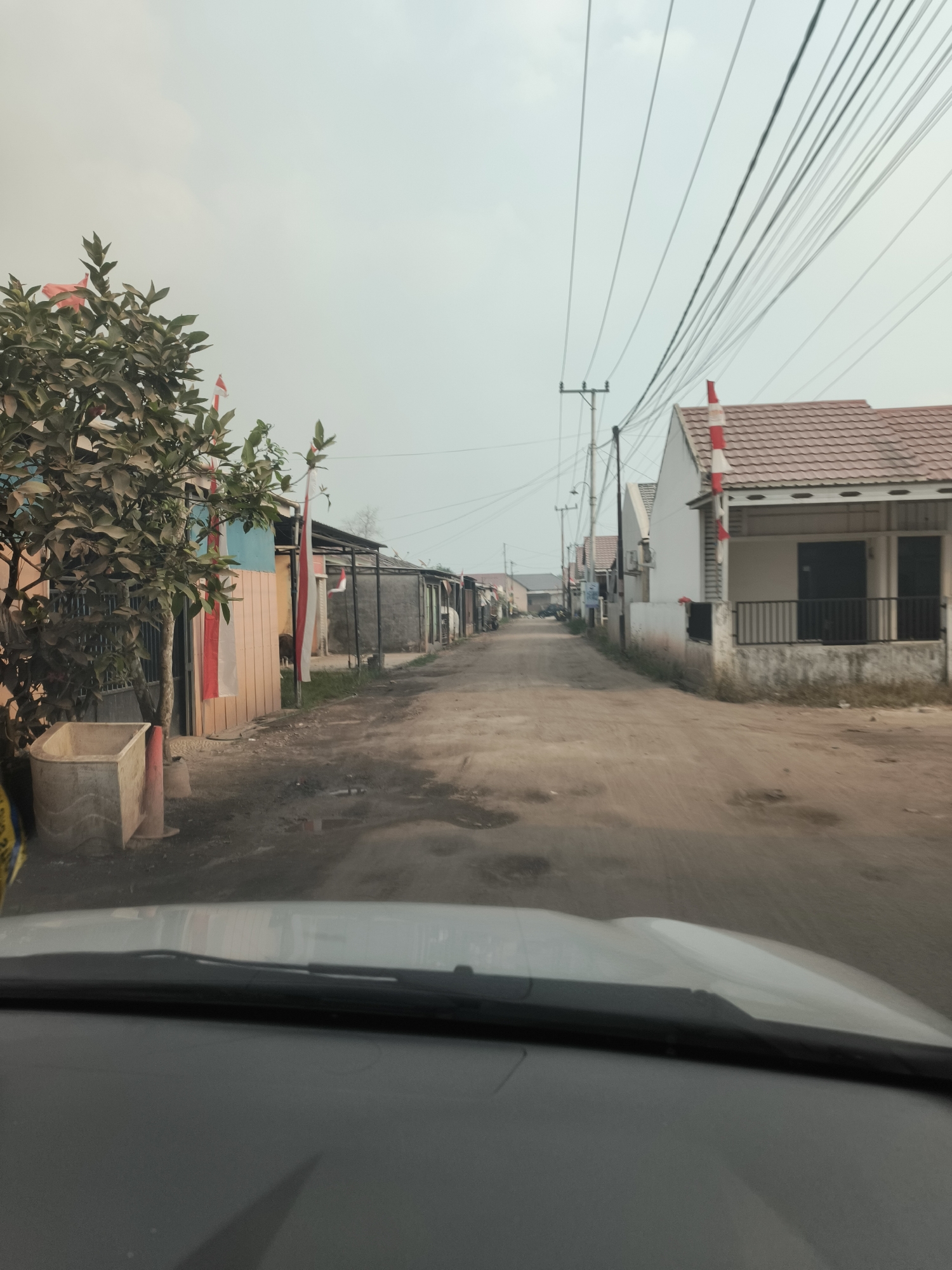 The image depicts a dusty, unpaved street in what appears to be a residential area in Sampit, Indonesia. The sky is overcast and a hazy white. On the left side of the street, a tree with green leaves is visible, with some red and white Indonesian flags attached to poles. Adjacent to the tree is a textured, light-colored rectangular structure, possibly a bin or planter. Further down the street on the left are several simple, narrow buildings with corrugated metal roofs and what appear to be colorful painted facades. Indonesian flags are also displayed in front of some of these buildings.
On the right side of the street, a row of more modern, white-painted houses with red tile roofs is present. These houses have covered porches with railings. Several utility poles with numerous electrical wires stretch across the upper portion of the image, dominating the sky. The road surface is dirt and shows signs of wear and scattered puddles. The perspective suggests the photo was taken from inside a vehicle, looking forward, with the dashboard visible in the foreground. The overall impression is one of a developing or somewhat modest urban or suburban environment.