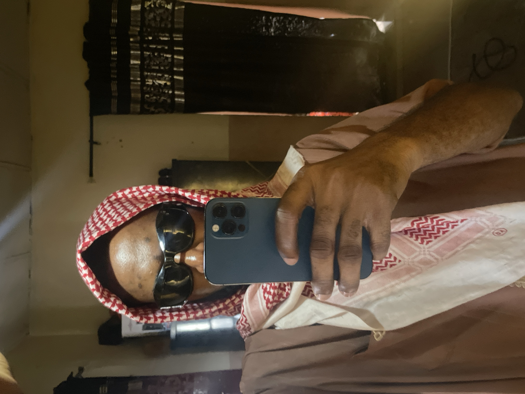 A person wearing a red and white checkered keffiyeh and sunglasses is holding a blue smartphone up to a mirror. The mirror's reflection shows a dark-colored textile hanging on a wall. The location in Lokoja, Nigeria, cannot be determined from the image.