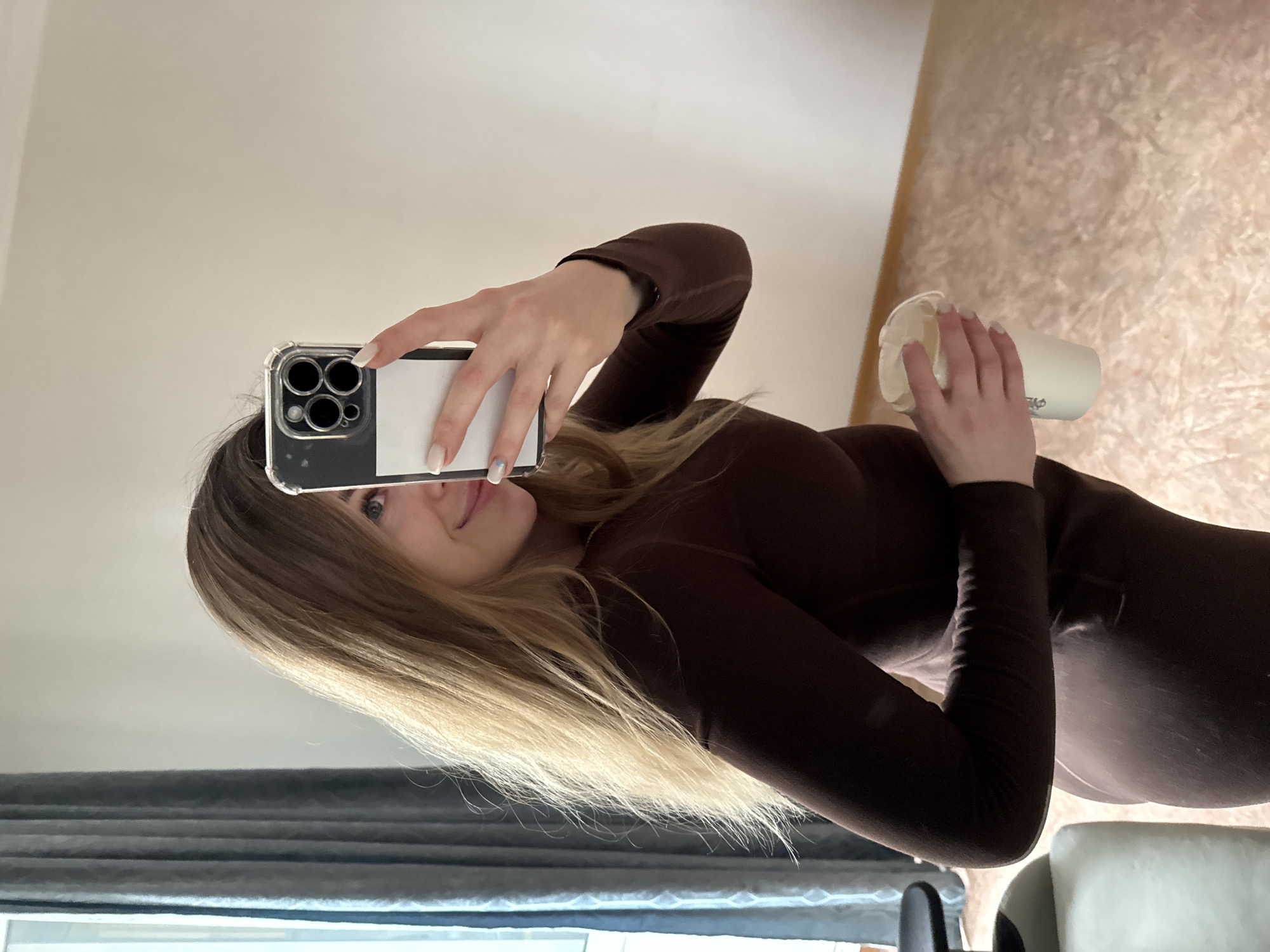 A person with fair skin and blonde hair is taking a selfie with a smartphone in a clear case, holding the phone in their right hand, obscuring the left side of their face. They are wearing a long-sleeved brown dress. Their left hand is holding a white cup with a lid. The background is composed of a light-colored wall, a window with gray curtains, and a carpet. The location appears to be indoors, possibly a residence. The city and specific location are Unknown, Unknown.