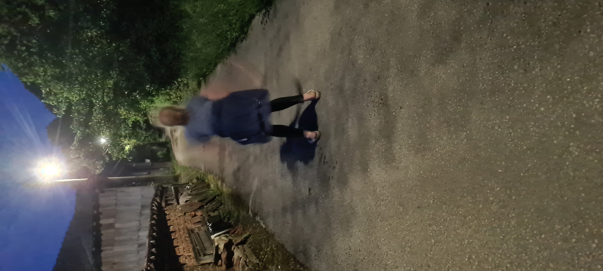 The image depicts a nighttime scene on a paved road in Lopian, Bulgaria. An adult individual, viewed from behind, stands centrally on the road. The person has light-colored hair and is wearing a light-colored top, dark jacket, black leggings, and light-colored sandals. Their posture is slightly bent forward, and there is a visible blur around the body, indicating movement during the exposure. A distinct shadow is cast on the road surface directly in front of the individual.
To the left of the road, an older building is visible, featuring a lower section of reddish-brown brick or stone and an upper section clad in light-colored, possibly corrugated, material. Debris or stacked items are accumulated against the brick base. A bright street lamp mounted on a pole stands adjacent to this building, casting strong illumination and creating a radial glare. Further down the road, to the right of this primary light source, a second, fainter light can be seen among dense green foliage.
The right side of the road is bordered by a strip of grass and extensive, dark green trees and bushes, suggesting a natural or unmanaged area. The road itself is a grey, asphalt-like surface with a coarse texture. The sky above is a deep blue, indicative of dusk or night, with subtle streaks of light cloud formation visible in the upper right quadrant. The overall environment suggests a rural or village setting.