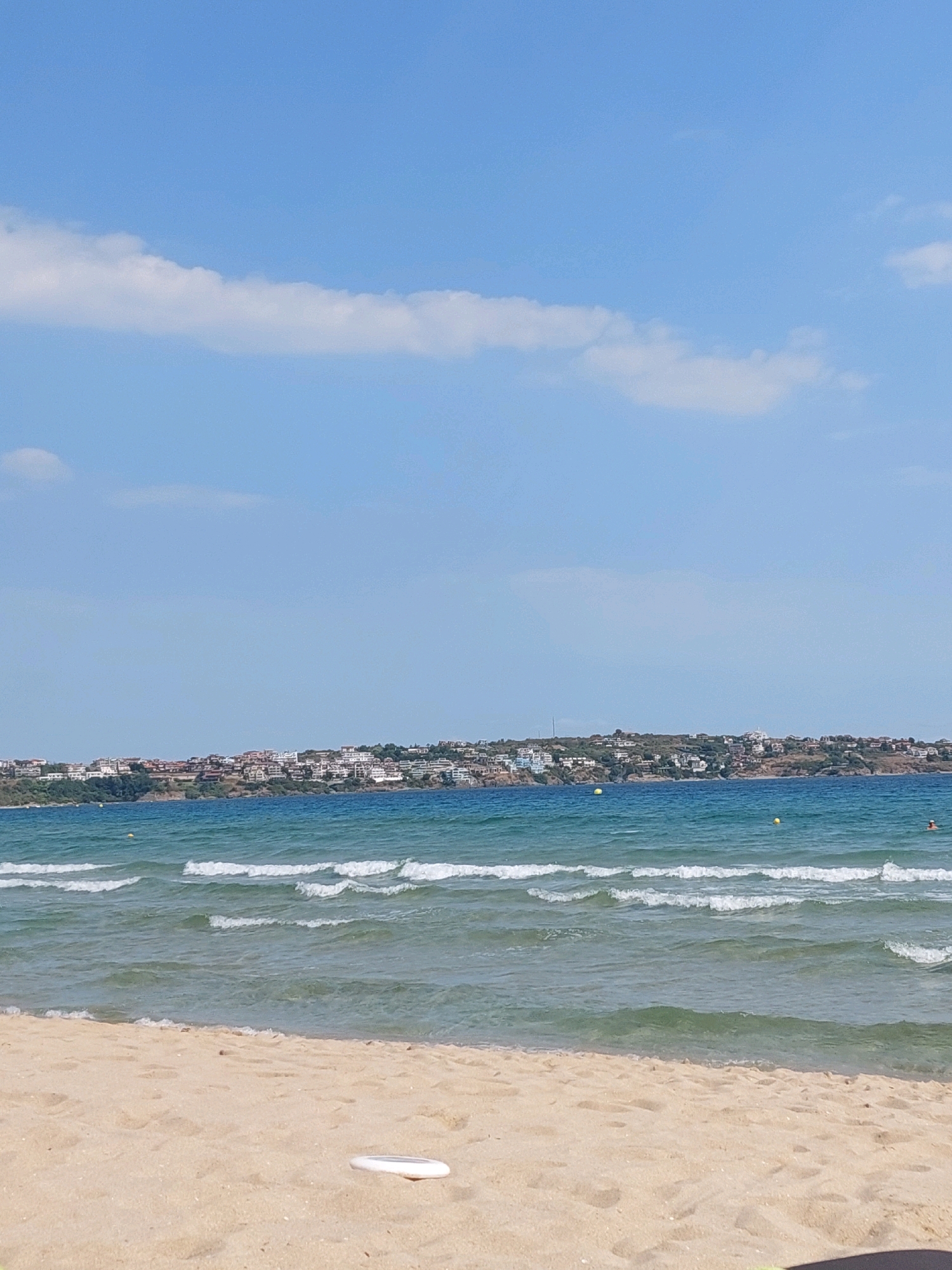 The image depicts a serene beach scene in Sozopol, Bulgaria. In the foreground, a stretch of sandy beach meets the gentle waves of the sea. A white frisbee rests on the sand. The middle ground is occupied by the turquoise sea with white-capped waves rolling towards the shore. In the distance, a coastline is dotted with buildings, indicating a town or resort area. The sky above is a clear blue with a few wispy white clouds. One person can be seen swimming in the sea near the distant shore, and two yellow buoys are visible in the water.