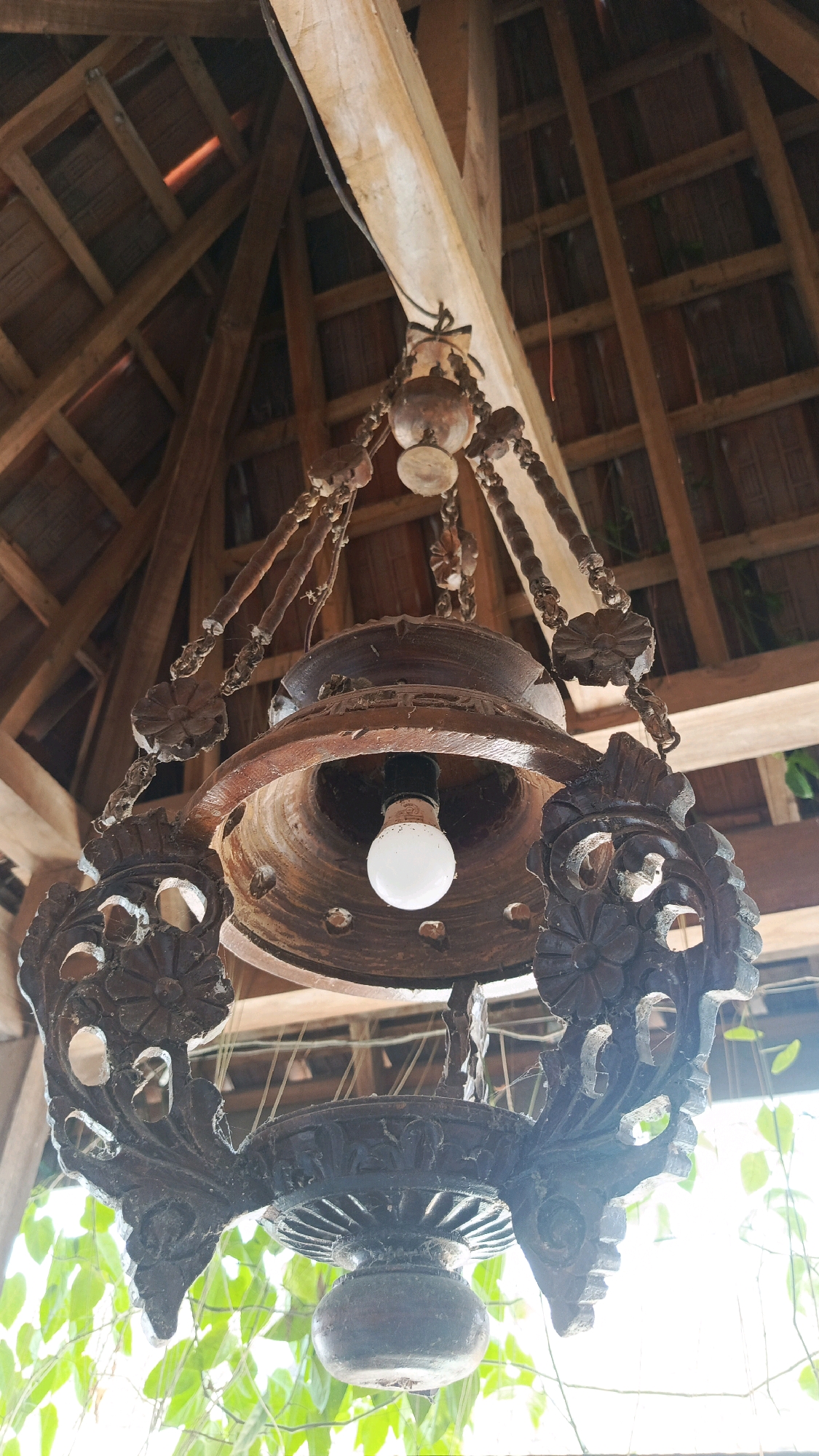 The image depicts a detailed, handcrafted wooden chandelier hanging from the wooden rafters of a building's roof. The chandelier is ornately carved with floral and scroll motifs. A single light bulb is visible inside the central housing. The background shows the structural elements of the roof, suggesting an indoor or covered outdoor space. Lush green foliage is visible at the bottom of the frame, indicating the presence of plants or trees outside the structure. The lighting suggests a bright, possibly natural light source filtering through the scene. The image was taken in Babadan, Indonesia.