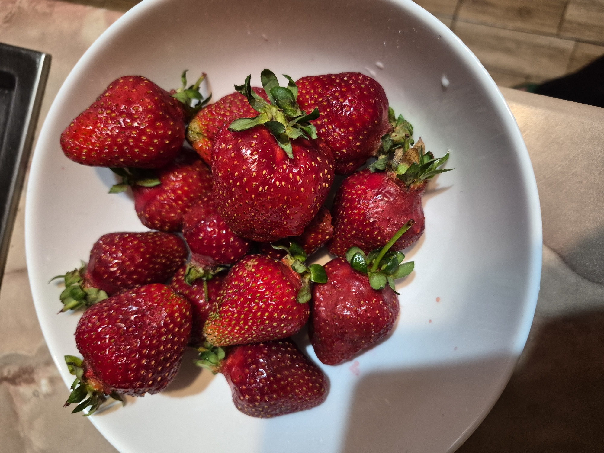 A close-up, high-angle shot captures a white bowl filled with numerous fresh, red strawberries. The strawberries are the primary subject, appearing plump and ripe, with their green calyxes still attached. Many have visible water droplets on their surface, suggesting they have been recently washed. Their vibrant red color is prominent, with some displaying slight variations in hue, including hints of yellow-orange near the top of a few berries. The achenes (seeds) are clearly visible as small, yellow dots embedded in the fruit.
The white, ceramic bowl holds a generous portion of the fruit, with some strawberries piled on top of others. The edges of the bowl are visible, creating a clean frame for the fruit.
In the background, to the left, a metallic edge, possibly part of a sink or counter, is partially visible. To the right and behind the bowl, a warm-toned surface with a subtle grain, resembling wood or a textured countertop, can be seen. The lighting appears to be artificial and warm, casting some soft shadows and highlighting the glistening berries. There is no discernible activity taking place other than the static arrangement of the fruit. No text is visible in the image. The setting appears to be an indoor kitchen or dining area, likely in Yerevan, Armenia.