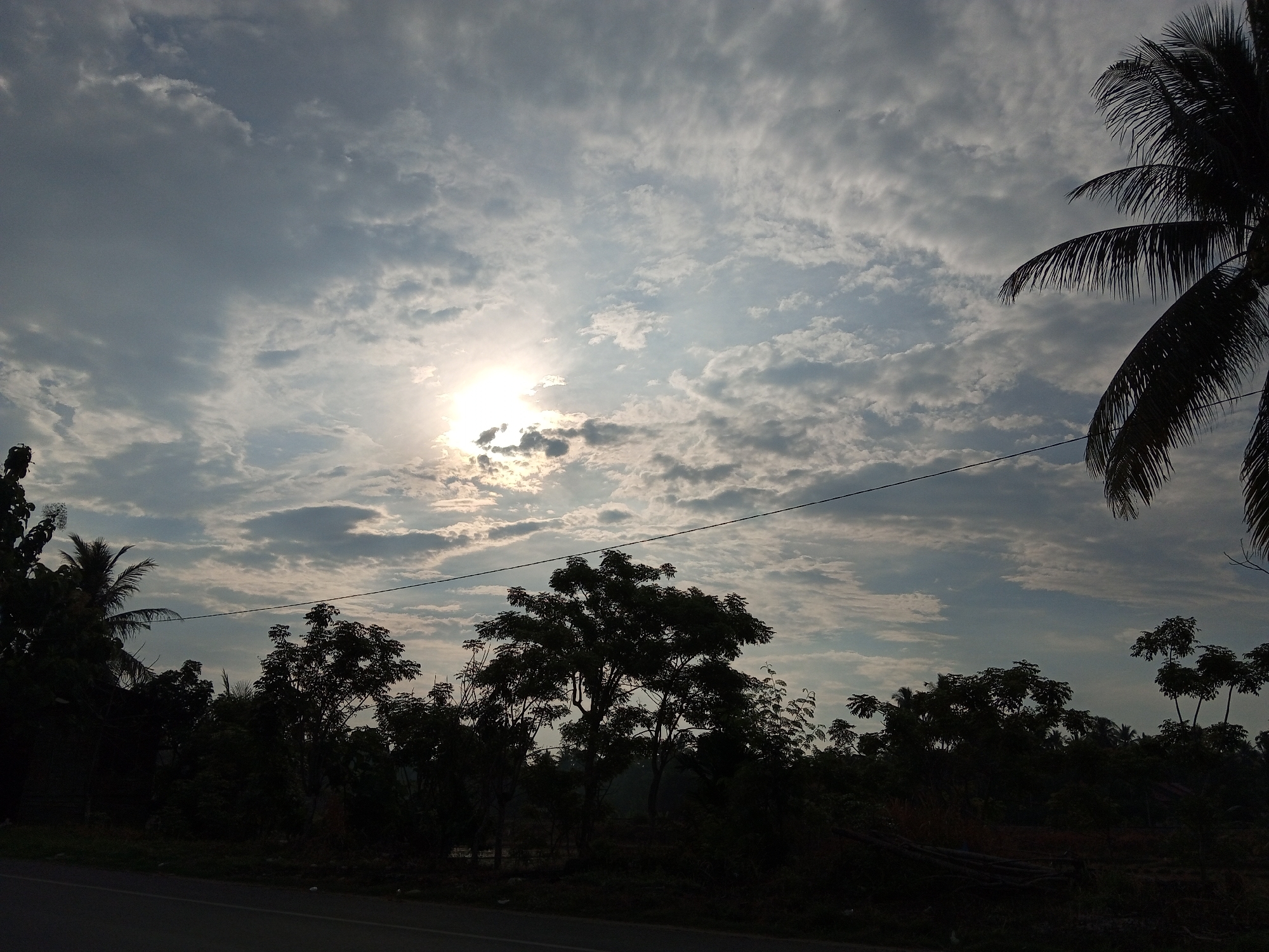 A view of the sky over Cotpuuk, Indonesia, features a bright sun peeking through scattered clouds. The lower portion of the image shows silhouettes of trees and foliage against the sky, with a power line stretching horizontally across the frame. A paved road is visible in the foreground. The scene appears to be captured during daytime, possibly early morning or late afternoon due to the low angle of the sun and the overall lighting.