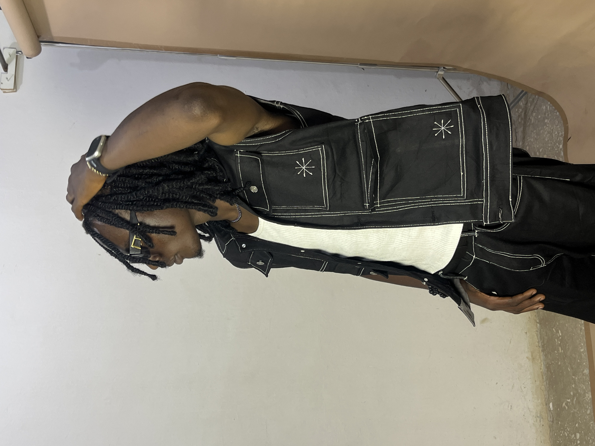 A person with dark skin and dreadlocks is shown from the side, leaning against a light-colored wall. They are wearing a white tank top under a black vest with white stitching. The vest has four pockets, two on the chest and two lower down, each with a small embroidered star. A silver watch is visible on their left wrist, and they are wearing glasses. The person has their left arm raised, with their hand resting on the back of their head. The scene appears to be indoors, with a glimpse of a light-colored ceiling and a beige-colored overhead structure. The lighting suggests it is daytime. There is no visible text.
