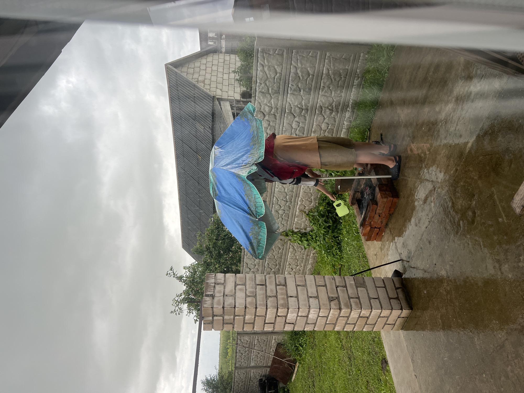 In Бучацінскі сельскі Савет, Belarus, on a rainy day, two individuals stand under a blue and green umbrella near a brick barbecue. One person, wearing a red garment over a white shirt and black pants, holds a green watering can, appearing to pour water onto the barbecue coals. The other individual, dressed in a tan shirt and beige shorts, stands beside them. The ground is wet, reflecting the overcast sky. A partially constructed brick pillar stands to the left, and a concrete fence forms the background. Behind the fence, a house with a grey roof and a grassy area are visible.