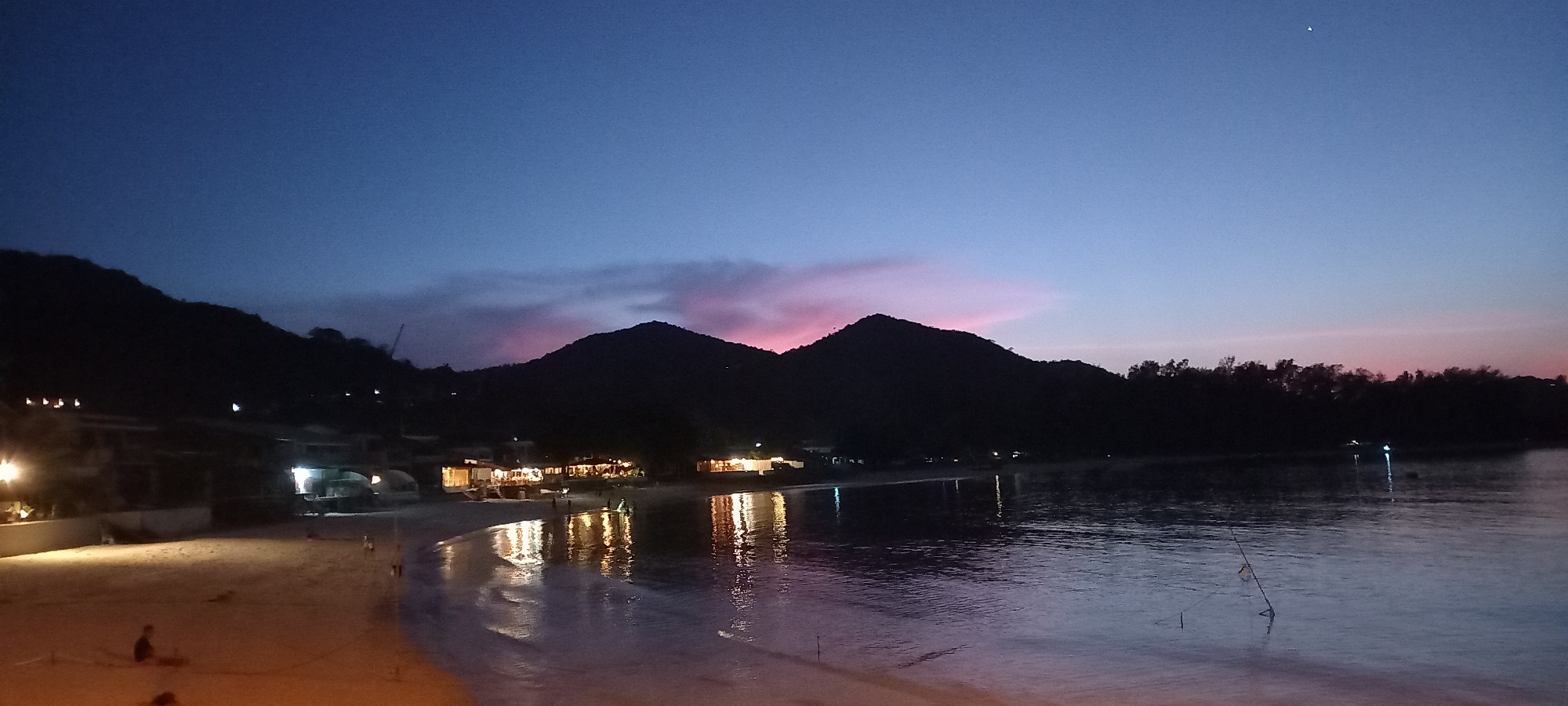 The image captures a serene coastal scene at twilight, showcasing a wide bay or inlet at Baan Chaloklum, Thailand. Dark silhouettes of mountains or large hills dominate the background, stretching across the horizon. Above them, the sky displays a gradient of colors, transitioning from a deep indigo-blue overhead to lighter blues and vibrant pink and purple hues closer to the horizon, indicative of the lingering glow of sunset. A faint star or distant light source is visible high in the sky towards the right.
Along the left side, a sandy beach is illuminated by warm artificial lights emanating from several buildings, likely restaurants or resorts, nestled against the mountainside. These lights cast bright, shimmering reflections onto the calm water of the bay. The water itself also reflects the soft colors of the twilight sky.
A few indistinct figures, appearing as dark silhouettes, are visible on the illuminated beach and at the water's edge; one person appears to be seated on the sand in the bottom left. On the right side of the water, a few thin poles or sticks protrude from the surface. In the background, behind the left mountain, a faint outline of a construction crane can be discerned. The scene evokes a tranquil and relaxed evening ambiance by the sea. No visible text is present.