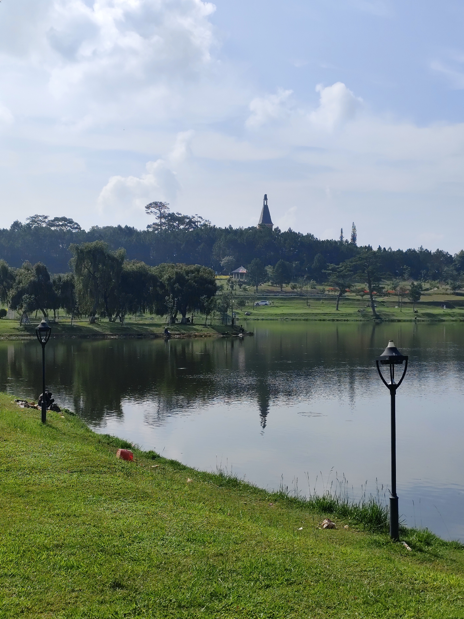 This image presents a picturesque lakeside park scene in Da Lat, Vietnam. The foreground is dominated by vibrant green grass, sloping gently towards a calm lake. Two dark, ornamental lampposts are prominent, one on the left and another on the right, framing the water's edge. Near the left lamppost, a red bucket or container is visible on the grass, and a person is seated on the bank, appearing to be fishing or relaxing by the water.
The still surface of the lake beautifully reflects the bright, partially cloudy sky and the trees lining the opposite bank. This far bank features a variety of lush green trees, some with distinctive weeping branches, and a dense forest that covers a rising hillside in the background. On this distant hill, a notable building with a dark, tall, and pointed spire or roof stands out against the sky, resembling a bell tower or chateau. A white vehicle and a small, gazebo-like structure can also be seen on the far bank, amidst other individuals enjoying the park.
The weather appears pleasant, with a bright sky featuring large, fluffy white clouds, suggesting it is daytime, likely morning or early afternoon. The overall atmosphere conveys peace, tranquility, and a sense of leisure in a natural setting. No visible text is present in the image.
