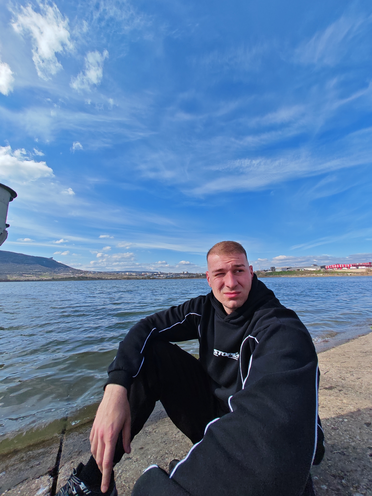 A young, light-skinned man sits on a concrete structure adjacent to a body of water, possibly a river or lake. He is wearing a black hoodie with white trim and black sweatpants, also with white trim. He has short, light brown hair. He is looking off to his right.
Behind him is a body of water, and in the distance, a low mountain range and cityscape are visible. The sky is a bright blue with streaky clouds. The location is near the Tombl Bayar Bridge in the city of Shumen, Bulgaria.