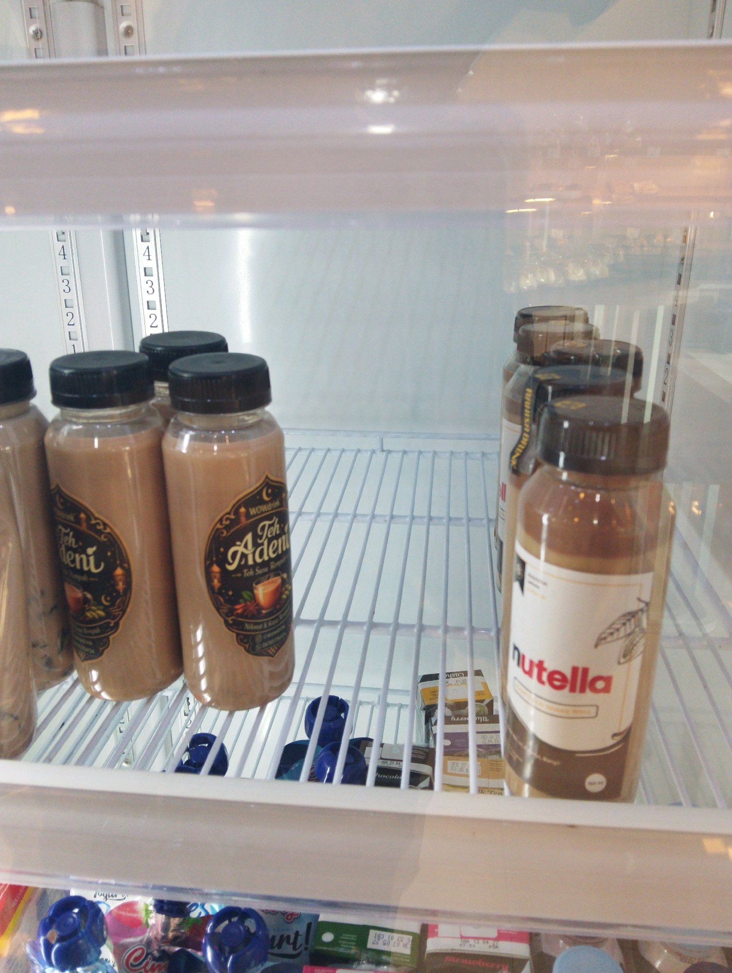 This image shows the inside of a refrigerated display case, likely in a convenience store in Manaruwi, Indonesia. The primary subjects are several bottles of beverages with brown liquid. On the left, there are at least four bottles with black caps and circular labels featuring text in what appears to be Malay or Indonesian, with decorative elements like crescent moons and lanterns. The labels also depict an image of a cup and some spices. To the right, there are more bottles, including one prominently labeled "Nutella" in red lettering, with a picture of a cacao pod and leaf. This bottle has a brown cap and a creamy brown liquid. Behind these bottles, other items are visible, including small cartons or boxes stacked on a lower shelf. The display case is well-lit, and the shelves are made of white wire racks. The background appears to be the interior of the refrigerator, with a white, ribbed texture. The overall impression is of a chilled beverage section in a retail setting.