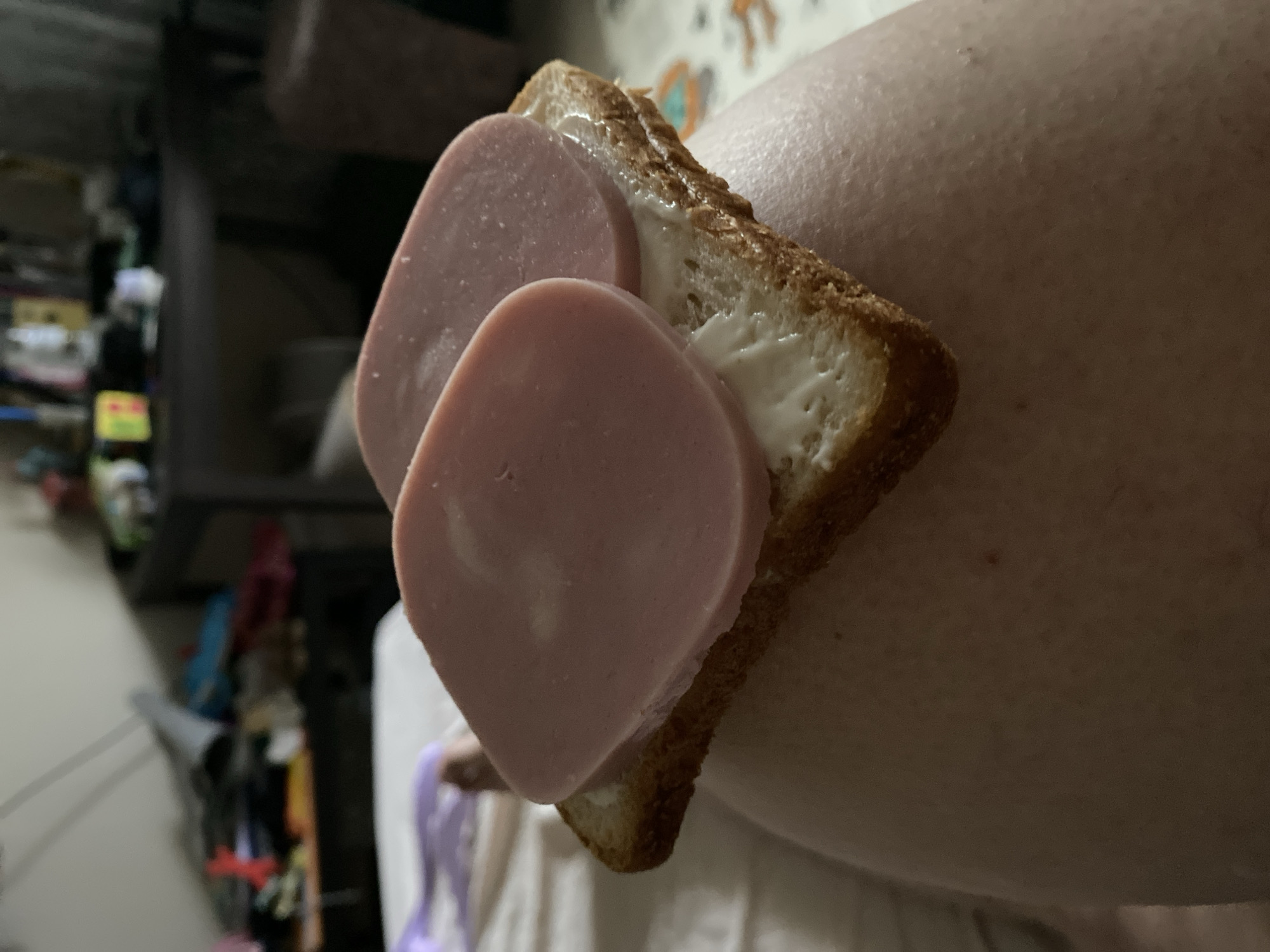 A close-up, low-angle shot shows a slice of toast with mayonnaise and two slices of ham, resting on a person's lap. The background is blurred, suggesting an indoor setting, possibly a bedroom, with a dresser or table laden with items. The lighting is dim, casting shadows and highlighting the texture of the toast and ham. The location is Sergiyev Posad, Russia.
