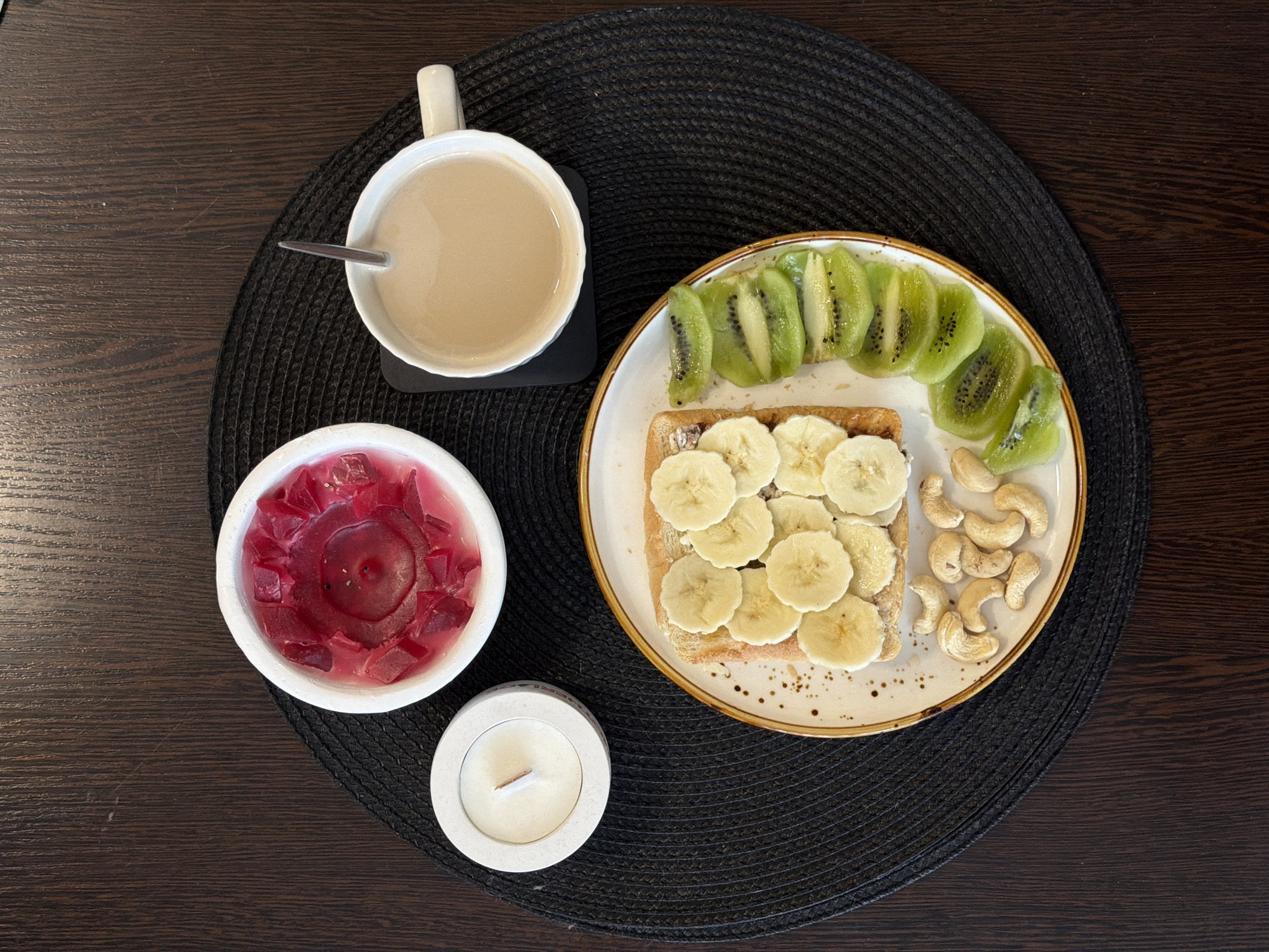 This top-down shot captures a calm breakfast or snack setting arranged on a dark brown wooden table. A round, black woven placemat serves as the base for the meal and decorative elements.
In the upper left quadrant, a white ceramic mug with a handle holds a light brown beverage, possibly coffee or tea with milk, with a spoon resting inside. Adjacent to it, on the right side of the placemat, a white ceramic plate with a textured brown rim and small brown speckles presents a balanced meal. The plate features a slice of toast or bread generously topped with neatly arranged slices of banana. To the right of the toast, a vibrant row of fresh kiwi slices, showcasing their green flesh and black seeds, adds color. Below the kiwi, a small pile of cashew nuts completes the food arrangement.
To the left of the plate, closer to the bottom, sits a white ceramic bowl containing a large, unlit red candle, possibly decorated with pieces of red wax or jelly-like fragments. Below this, a smaller, round, light-colored container, appearing to be made of concrete or ceramic, holds a small, unlit white candle with a wooden wick.
The scene suggests an indoor setting, likely a home, focusing on a personal moment of relaxation or a leisurely meal, possibly in the morning given the breakfast items. There are no visible people or text. The overall lighting is even, suggesting ambient indoor light. While the location context is given as Засечное, Russia, no specific visual cues in the image confirm this.