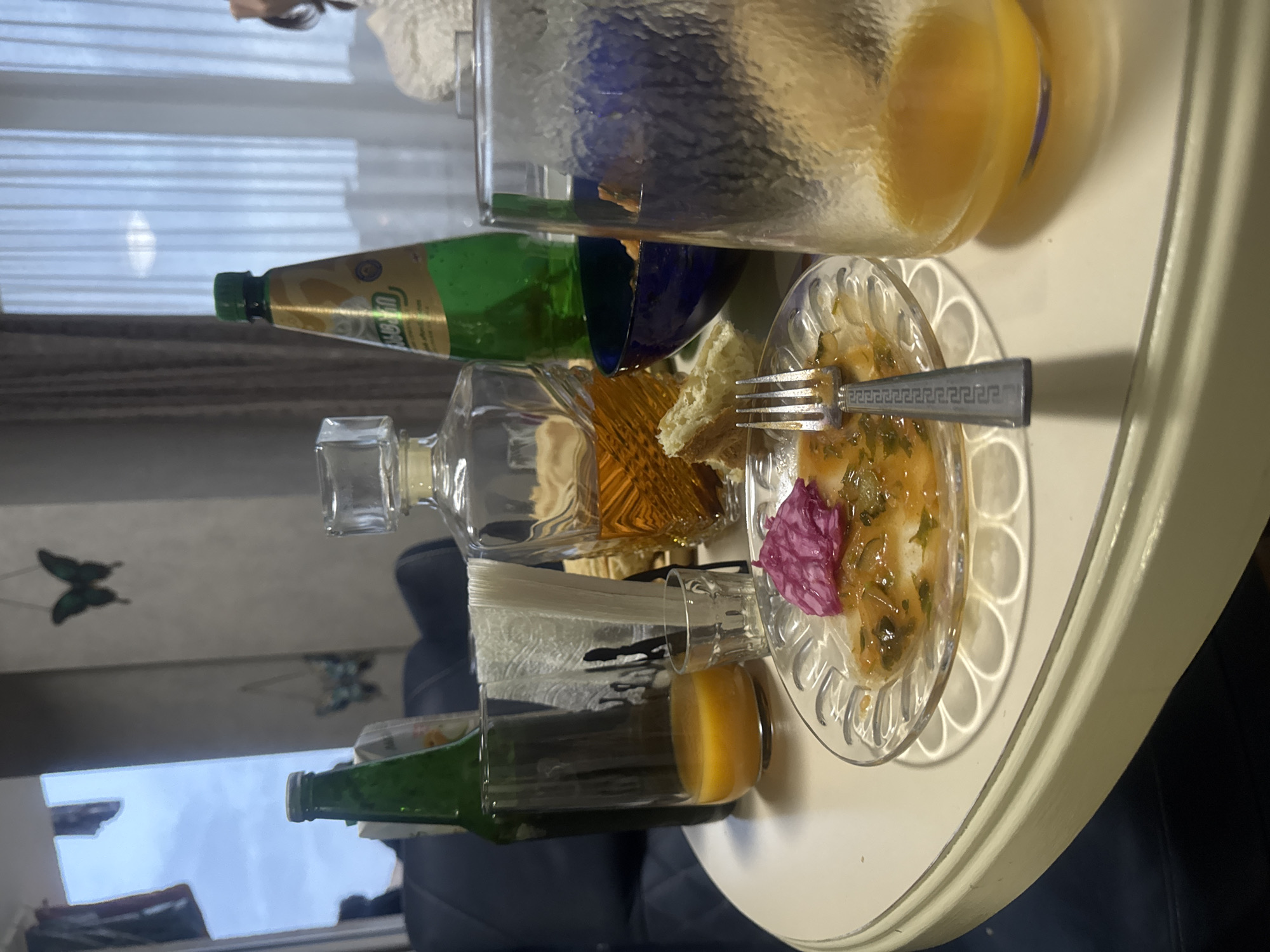 The image shows a cluttered table, likely in a residential setting based on the window with blinds and butterfly decorations in the background. Several objects are present on the table:
* **Liquids:** A large pitcher or jug filled with a yellow liquid (possibly juice), a bottle with a green label and green liquid, a glass bottle filled with an amber liquid, and a glass containing an orange liquid.
* **Food:** A clear plate with a dish that appears to be salad or a similar appetizer. The salad includes a pinkish-purple flower or vegetable element. Pieces of bread are also visible.
* **Utensil:** A silver fork is placed on the plate.
* **Other:** A stack of napkins is in a glass.
The scene is indoors. The image is slightly blurry, and the objects appear to be arranged somewhat haphazardly. While the general location is likely Tbilisi, Georgia (as requested), there is no visual information to confirm the exact address or public landmarks.